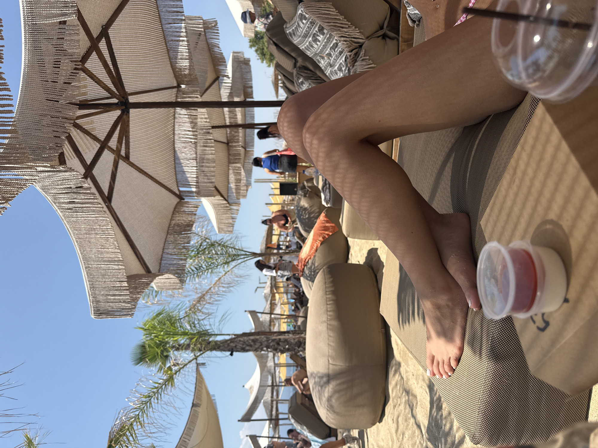 The image depicts a sunny day at Ofrynio Beach, Greece. In the foreground, a person's tanned legs are visible, resting on a brown lounge chair. A small plastic container with a red dessert or sauce and a clear plastic cup with a straw are placed on a cardboard surface in front of the chair. The background shows a beach scene with more lounge chairs, umbrellas with decorative fringe, palm trees, and other beachgoers in the distance. The sky is clear and blue.