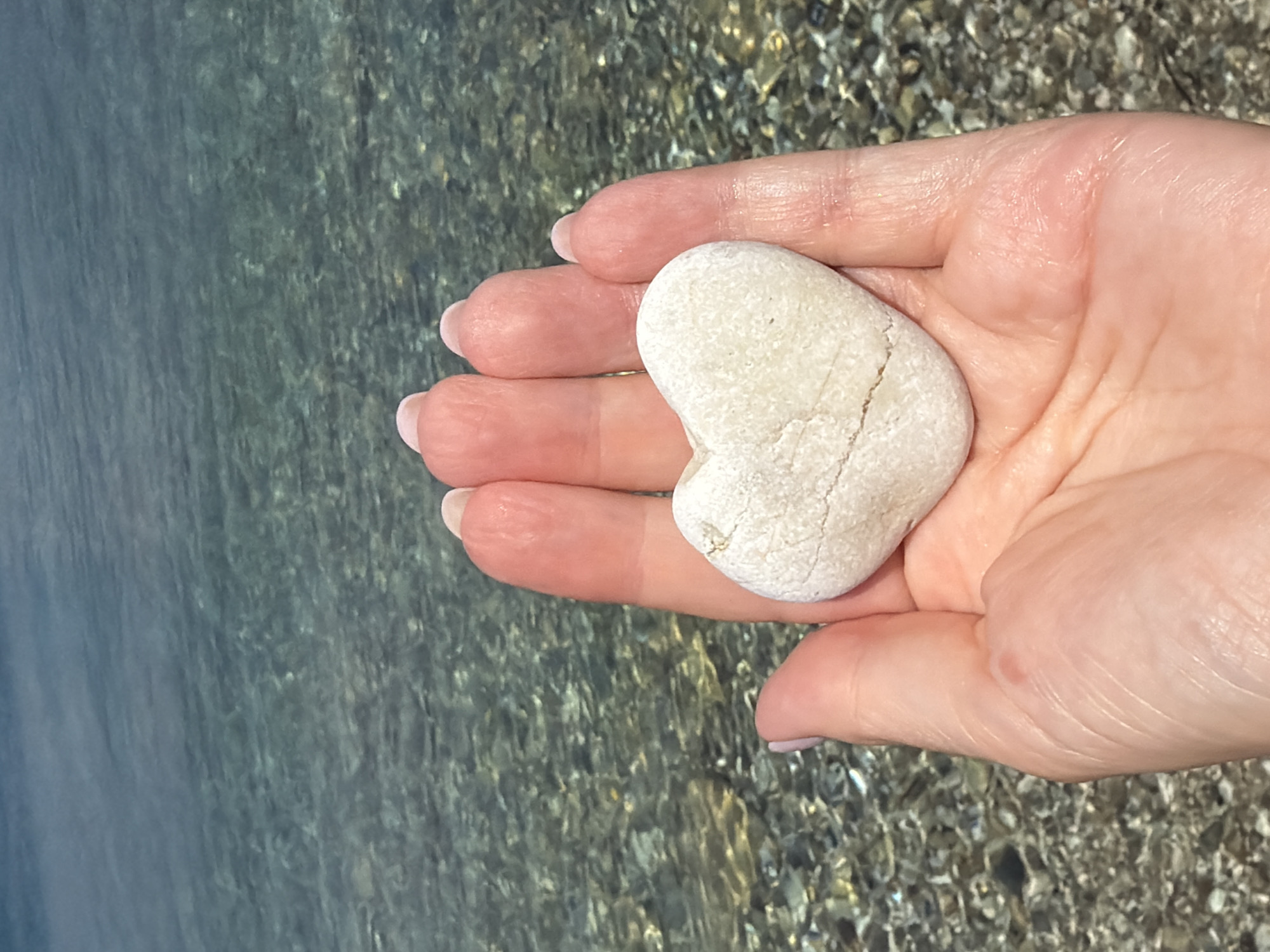 A close-up shot from a first-person perspective shows a pale, heart-shaped stone resting in a person's open palm. The hand, with its manicured fingernails, is positioned in the foreground, with fingers slightly curled. Behind the hand, the crystal-clear, shallow water of the sea creates a shimmering backdrop, revealing a rocky seabed below. The image appears to be taken on a beach, possibly in the Municipal Unit of Kastos, Greece, given the typical coastal scenery of the region. The water is calm and reflects the light, creating a serene and tranquil atmosphere.