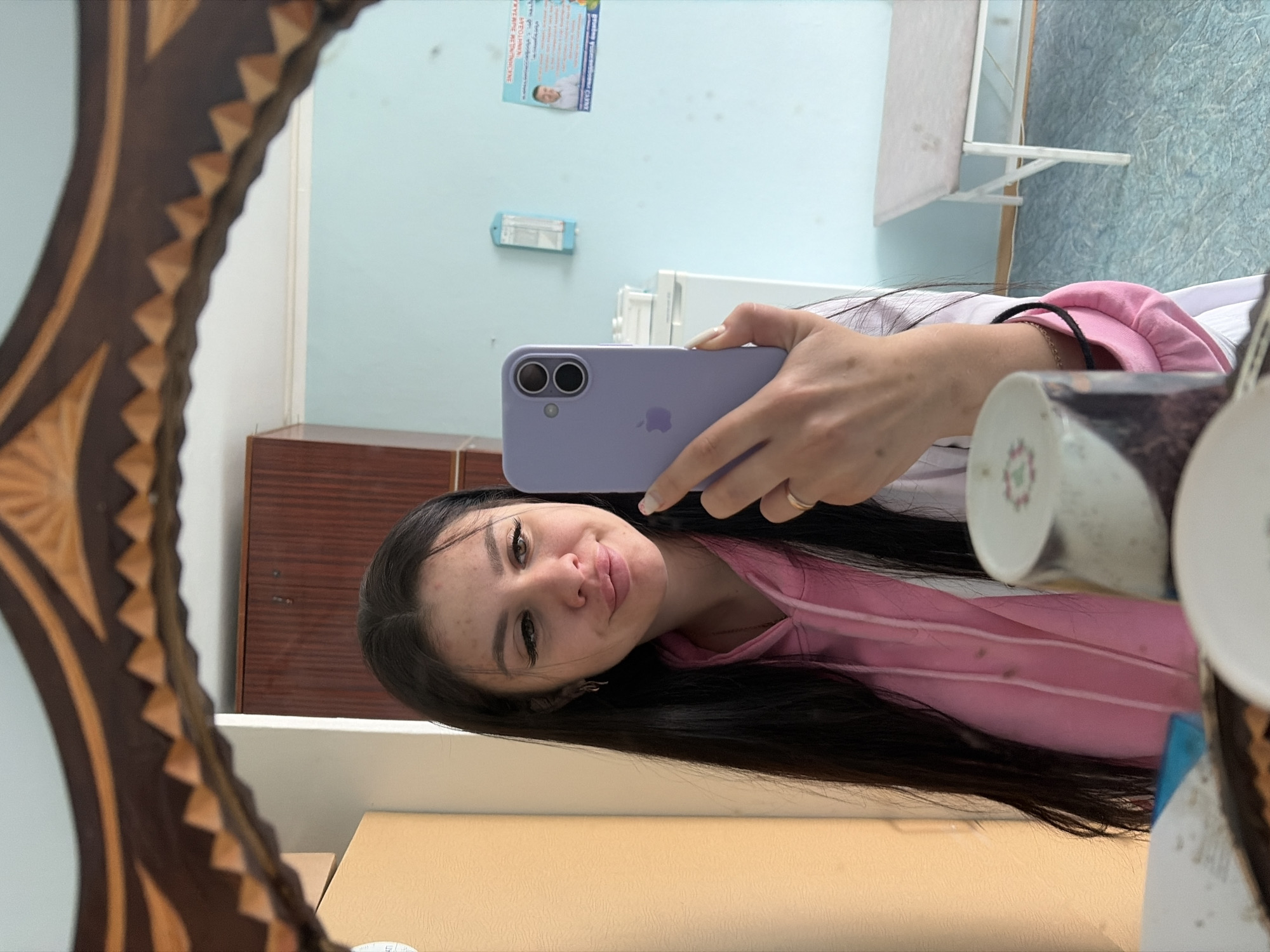 A young woman with long, dark hair takes a mirror selfie in what appears to be a room with light blue walls. She is wearing a pink hoodie and a white lab coat. She holds a lavender-colored smartphone in her right hand, capturing her reflection. The room contains a dark wood cabinet, a refrigerator, a bench with a light-colored top, and a poster on the wall. The floor is light blue with a textured pattern. A decorative wooden frame surrounds the mirror. The overall impression is that of a clinical or institutional setting, potentially a medical facility or dormitory, located in the city of Chumna, Russia.