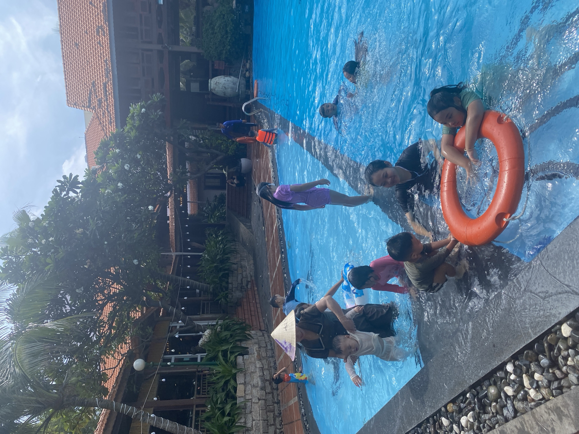 This image depicts a vibrant scene at a swimming pool in Phu Thuy Ward, Vietnam. Several children and adults are enjoying the water, with the pool water being a bright, clear blue. In the foreground, an adult wearing a conical hat is assisting a young child in the pool, while another child in a pink swimsuit is near them. Further into the pool, several other children are visible, some playing and others swimming. There is an orange life preserver ring in the water. The background shows a building with a red tiled roof, surrounded by lush green trees and palm trees, suggesting a tropical or subtropical setting. The overall atmosphere is one of recreation and family fun.