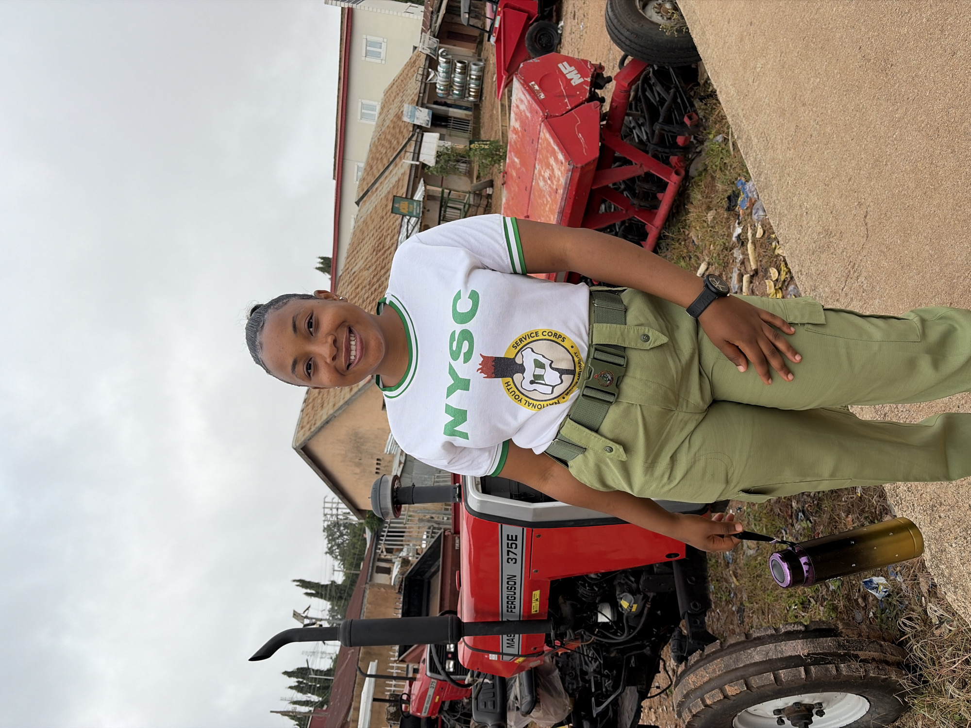 A young woman in a white NYSC t-shirt with green trim and olive green cargo pants stands in front of a red Massey Ferguson 375E tractor. She is smiling and holding a dark and gold water bottle in her left hand. The tractor is parked in an outdoor lot with several other pieces of red agricultural equipment visible in the background. Buildings with corrugated iron roofs are also visible behind the equipment. The ground is a mix of dirt, gravel, and some scattered debris. The sky is overcast. The location is in Jos, Nigeria.