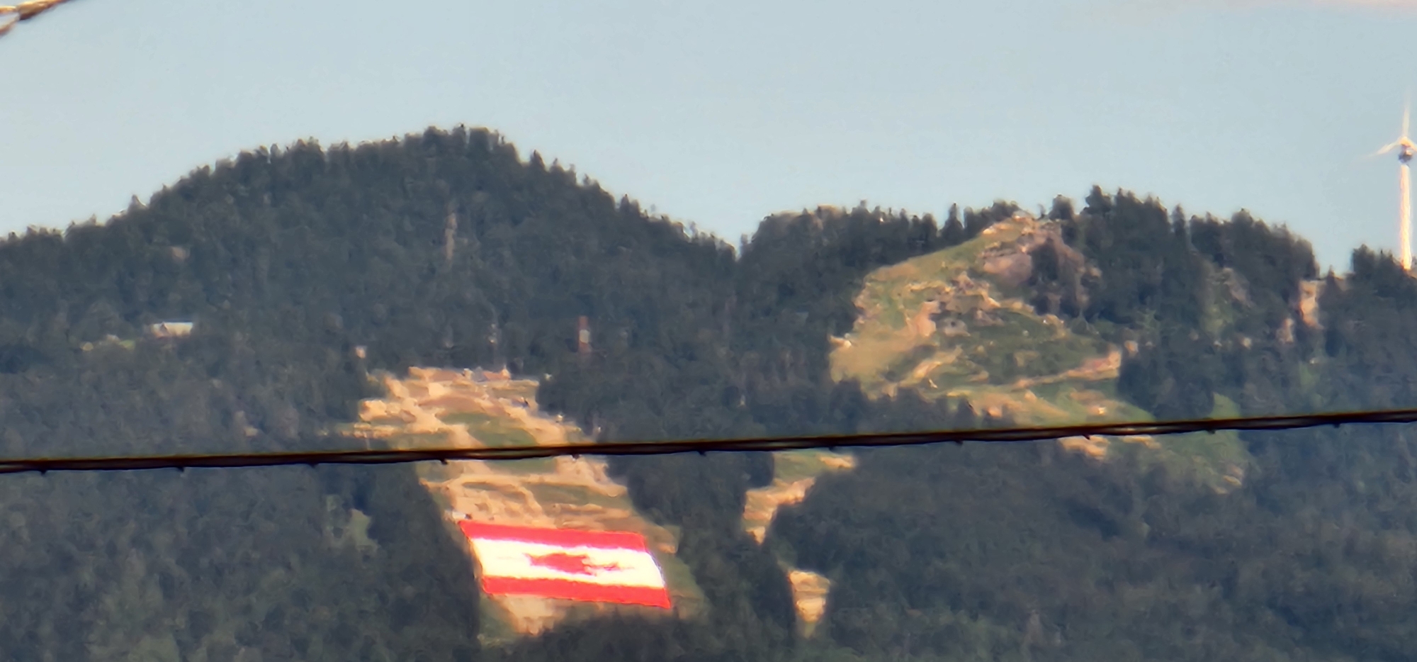 The image shows a wide, horizontal view of a mountain range covered in dark green trees. In the foreground, a dark, thin horizontal wire stretches across the frame. In the middle ground, a large, red and white flag is visible, appearing to be draped over a section of the mountain. The flag has a lighter colored symbol in the center. The mountain slopes show patches of lighter colored vegetation or cleared areas, with some shadows indicating uneven terrain. In the far background, a wind turbine is visible on the right side, and the sky is a clear, pale blue. The image appears to be taken from a distance, with a slightly blurry quality.