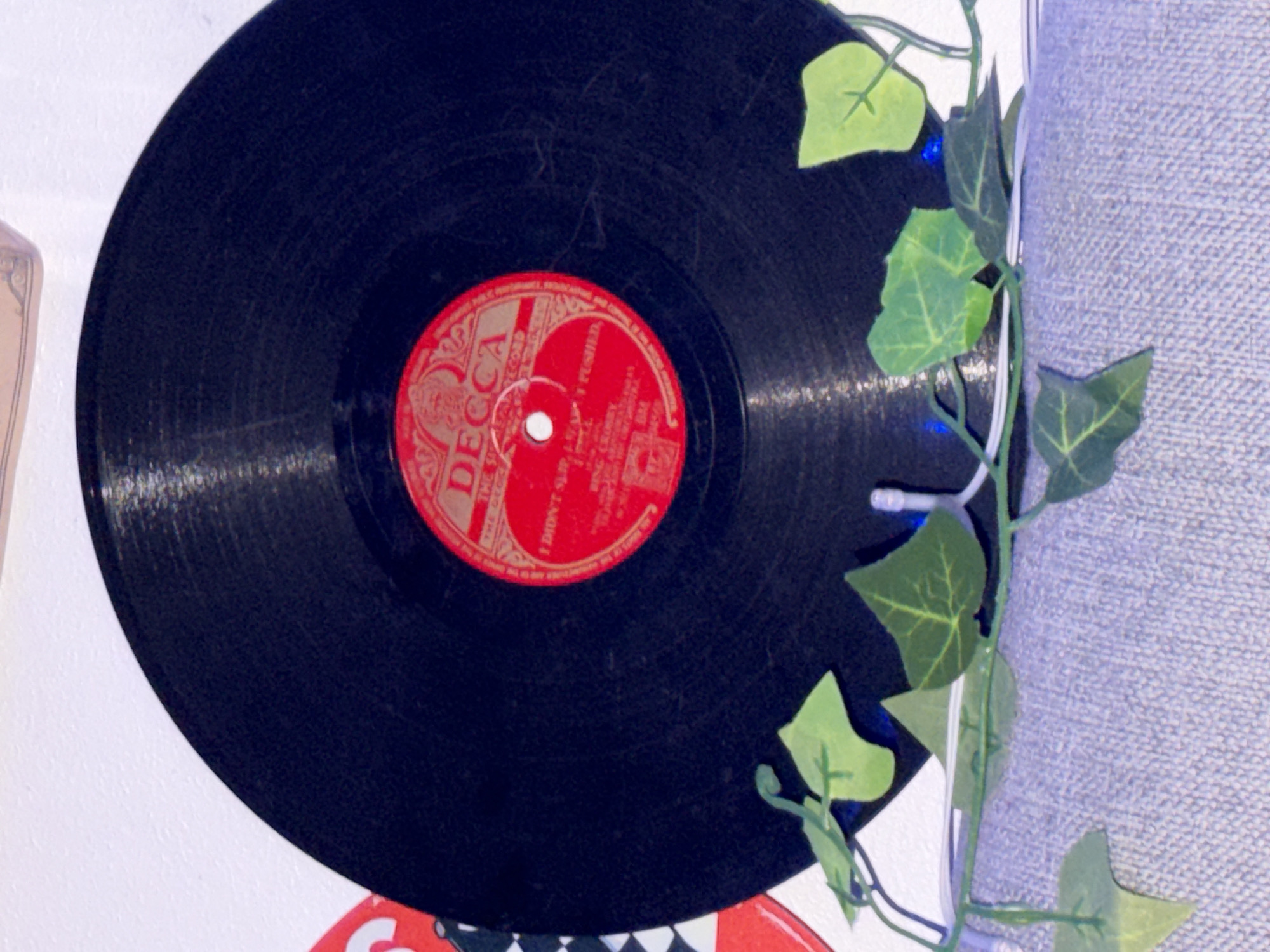 The image shows a close-up, overhead shot of a black vinyl record with a red and gold label. The label is circular and features the word "DECCA" prominently in large gold letters. There is text below "DECCA" that is difficult to read but appears to be "THE DECCA RECORD." Further down, smaller text indicates "I DIDN'T KNOW ABOUT YOU AND I PUSHED." Below that, it seems to list "BING CROSBY" and some other unreadable text. The record is positioned on what appears to be a textured grey surface, possibly a shelf or fabric. There is a string of artificial green ivy with small leaves draped across the bottom of the record, partially obscuring the grey surface. The background is a plain white wall. There is a hint of a checkered pattern on a red surface to the left of the record, suggesting it might be another record or a decorative item. The overall lighting is dim, with some blue light visible behind the ivy. There are no people or animals present in the image. The scene appears to be indoors, in a domestic or display setting. There is no indication of the location being in Oslo, Norway.