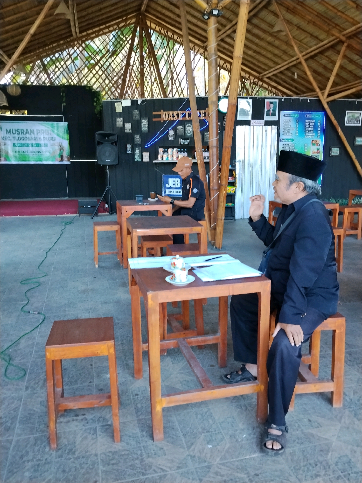 A middle-aged man in a black peci and dark blue shirt sits at a wooden table, gesturing with his right hand. He is in an outdoor cafe with a thatched roof and bamboo pillars. The cafe has multiple wooden tables and stools, and a stage with a banner in the background. Another man in an orange cap and black shirt sits at a table in the background. The floor is tiled and there are various decorations on the wall, including framed pictures and posters. The overall atmosphere suggests a casual gathering or event.