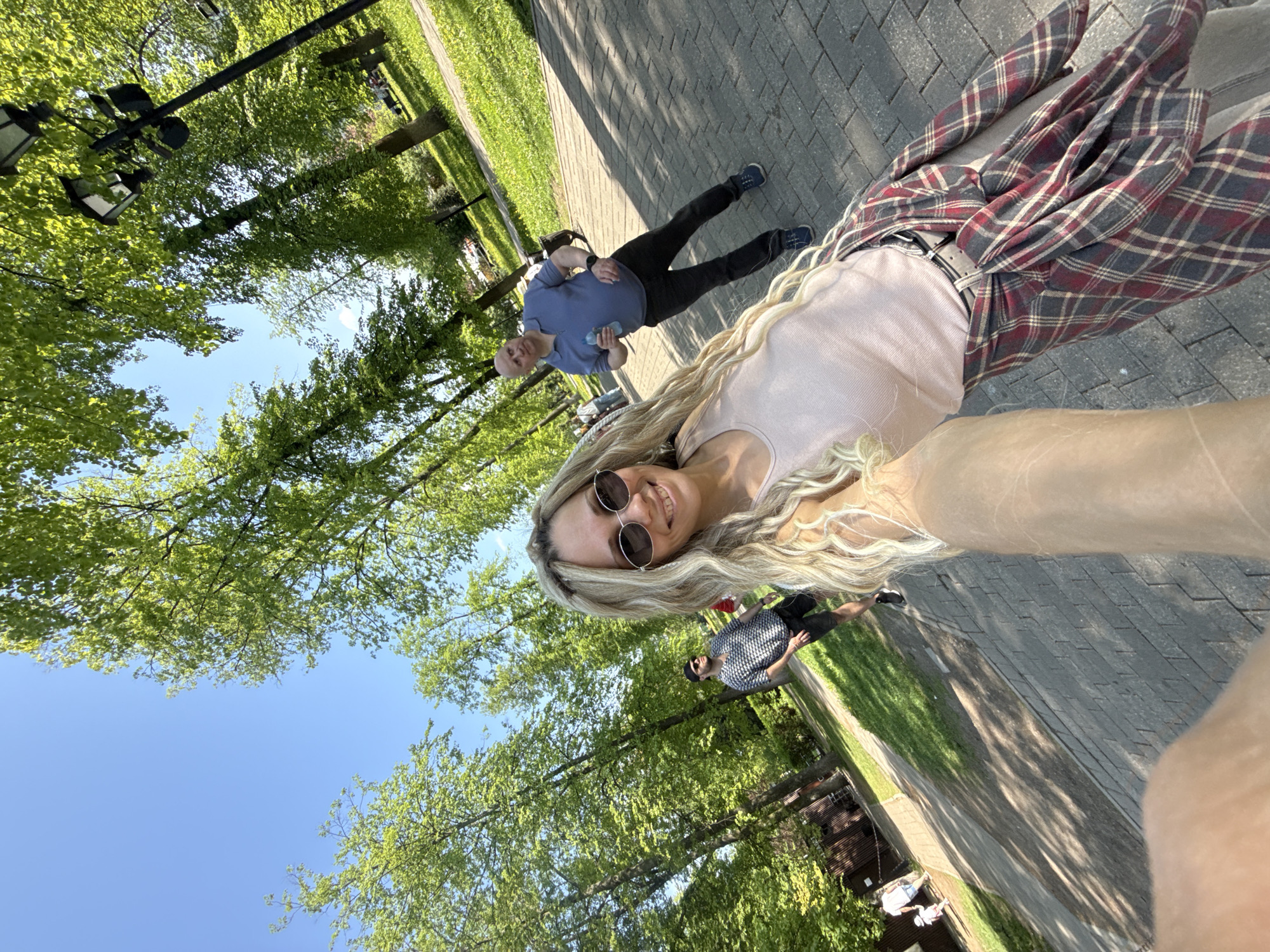 A sideways low-angle photograph depicts a woman in sunglasses taking a selfie on a brick walkway, in a park or urban setting. The woman has long, blonde, wavy hair and is wearing a light pink top and a flannel shirt tied around her waist. Two other men appear in the background. The first man is wearing a blue shirt and dark pants and holding a water bottle. The other man is wearing a black and white shirt and black shorts. The trees surrounding the walkway are lush and green, and there is a blue sky visible through the foliage. A light post appears in the upper left corner of the photograph.