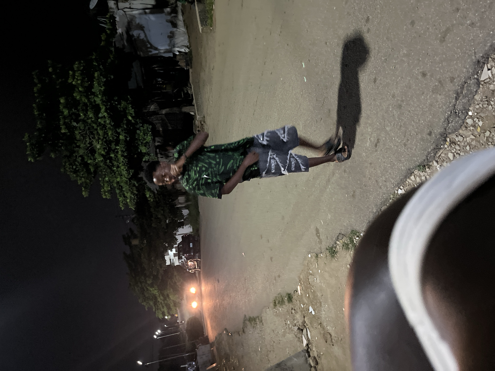 A full shot captured at night shows a young Black male standing on a paved road in a city. He is wearing a short-sleeved, green patterned shirt and distressed denim shorts with "GIVENCHY" text printed on them. He is also wearing black sandals. The male is looking to his right and is in the process of moving his right arm and hand towards his face. The road is lit by streetlights and the headlights of approaching vehicles, indicating an urban environment. The background shows trees and structures consistent with a residential or commercial area. The camera appears to be at a low angle, with a portion of another person's shoulder and arm in the foreground, out of focus. The overall scene suggests a casual moment outdoors at night.