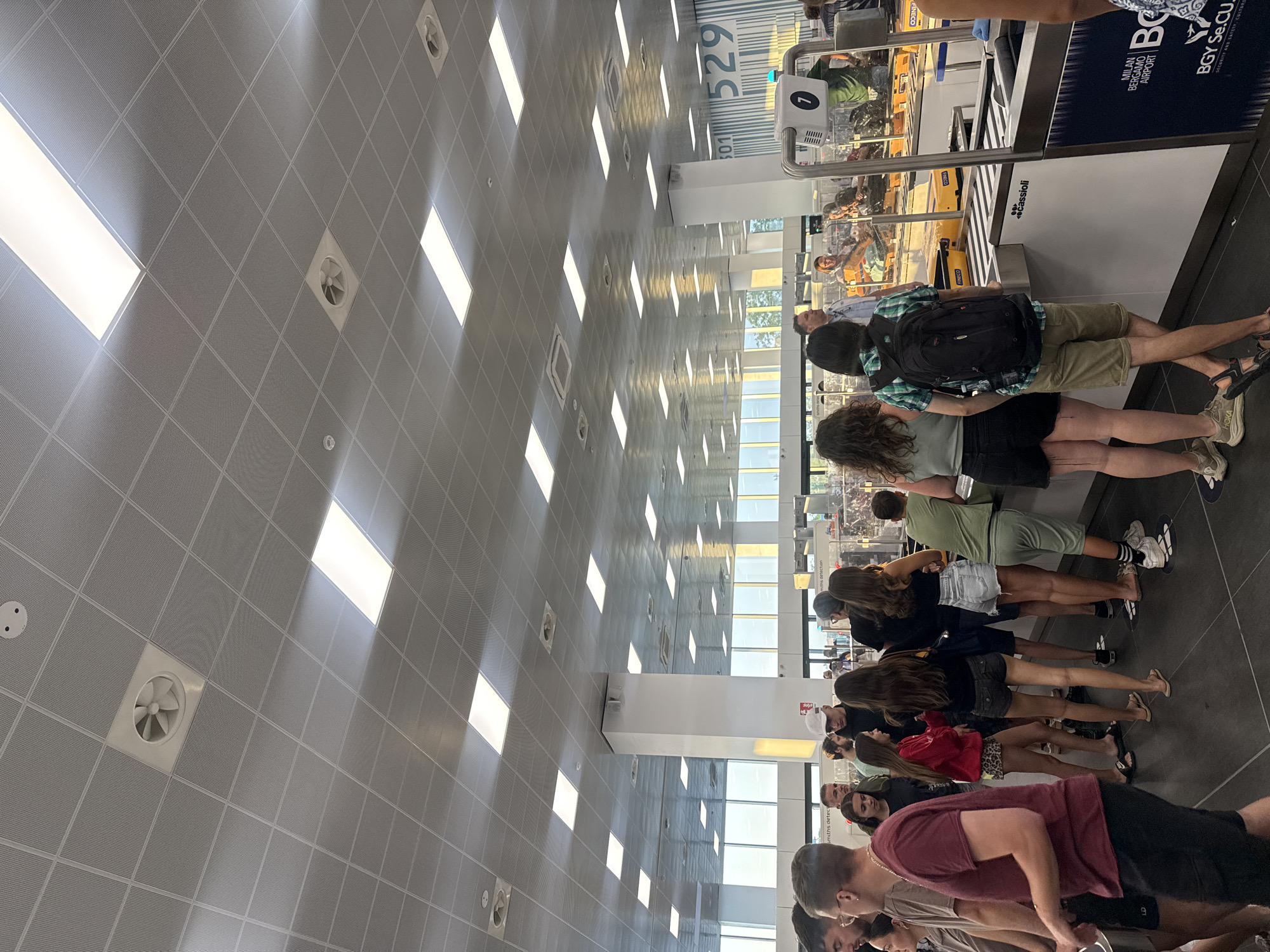 The image captures a scene inside Milan Bergamo Airport in Orio al Serio, Italy. A group of people, appearing to be travelers, are standing in line, likely for check-in or security. They are dressed casually in summer attire. The environment is brightly lit by fluorescent lights on the suspended ceiling, which also features ventilation grilles. In the background, luggage belts and security check points are visible, along with signage indicating "MILAN BERGAMO AIRPORT" and a code "529". The overall atmosphere suggests a busy travel hub.