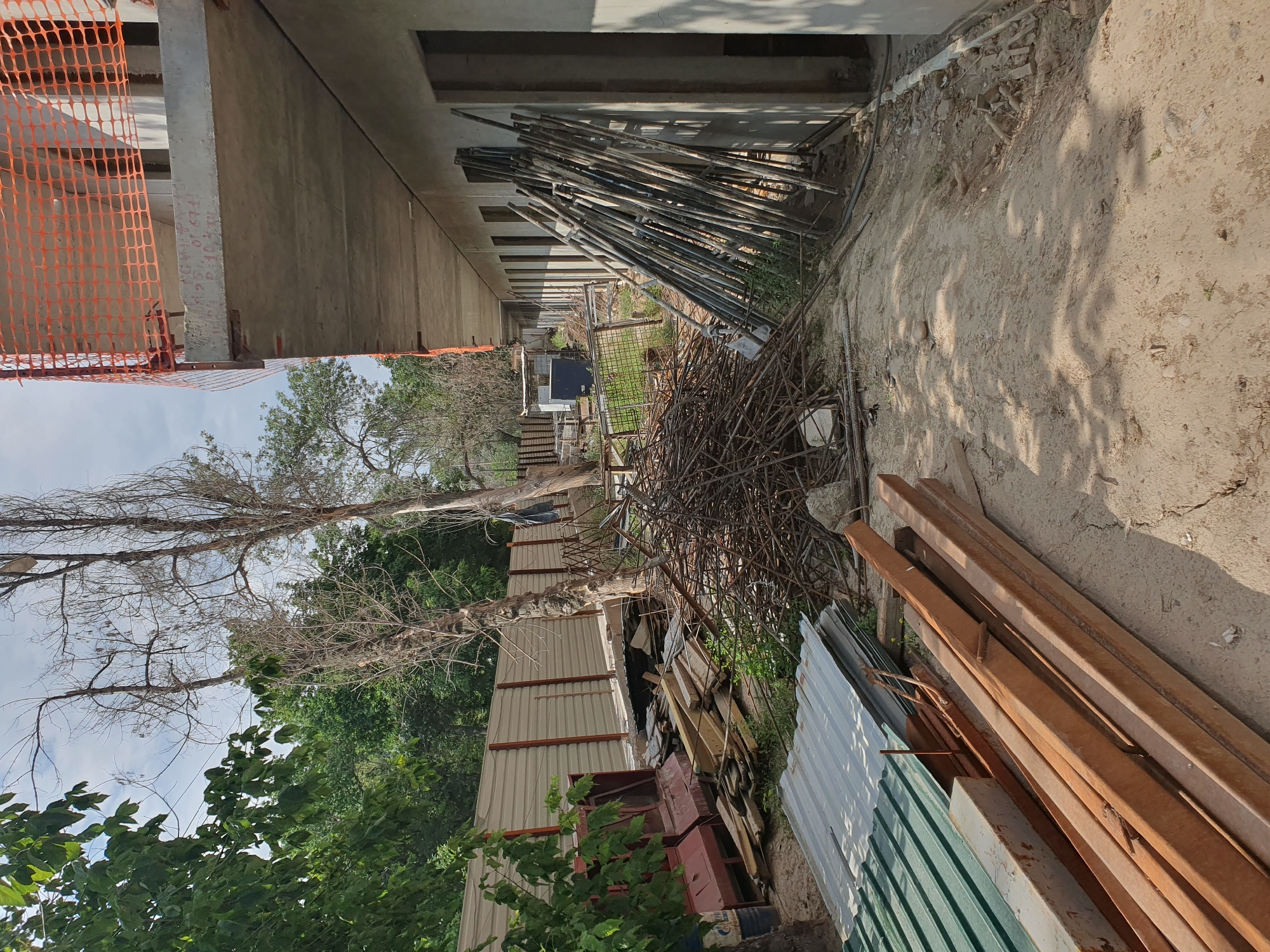 The image depicts an outdoor construction site in Tashkent, Uzbekistan, with a partially built concrete structure dominating the right side. The building appears to be a multi-story facility, with exposed concrete floors and openings for windows or doorways. Orange safety netting is visible on the upper level. In front of the building, a pile of rusted rebar is haphazardly arranged, suggesting recent construction activity. Various construction materials, including stacks of corrugated metal sheets and wooden planks, are scattered across the ground. The environment is characterized by a dusty, unpaved surface with scattered debris. Several tall, dry trees with sparse foliage are present in the background, interspersed with green trees and a metal fence. The sky is partly cloudy. There are no people visible in the scene.