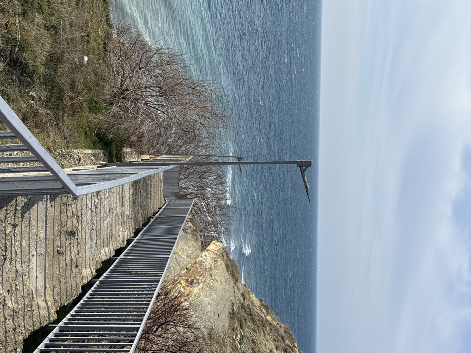 The image depicts a concrete staircase descending a coastal incline towards the Black Sea in Anapa, Russia. The staircase features silver-colored metal handrails on both sides and appears worn, with surface debris. To the left, a slope covered in sparse dry grass and leafless shrubs borders the steps. To the right, a multi-layered rocky cliff face with some vegetation parallels the descent. The choppy, deep blue sea extends to the horizon under a light blue sky with scattered white clouds. Two grey metal streetlights, each with a horizontal arm, are present; one stands prominently above the staircase, and a smaller one is partially visible further down. No individuals or active subjects are present in the scene.