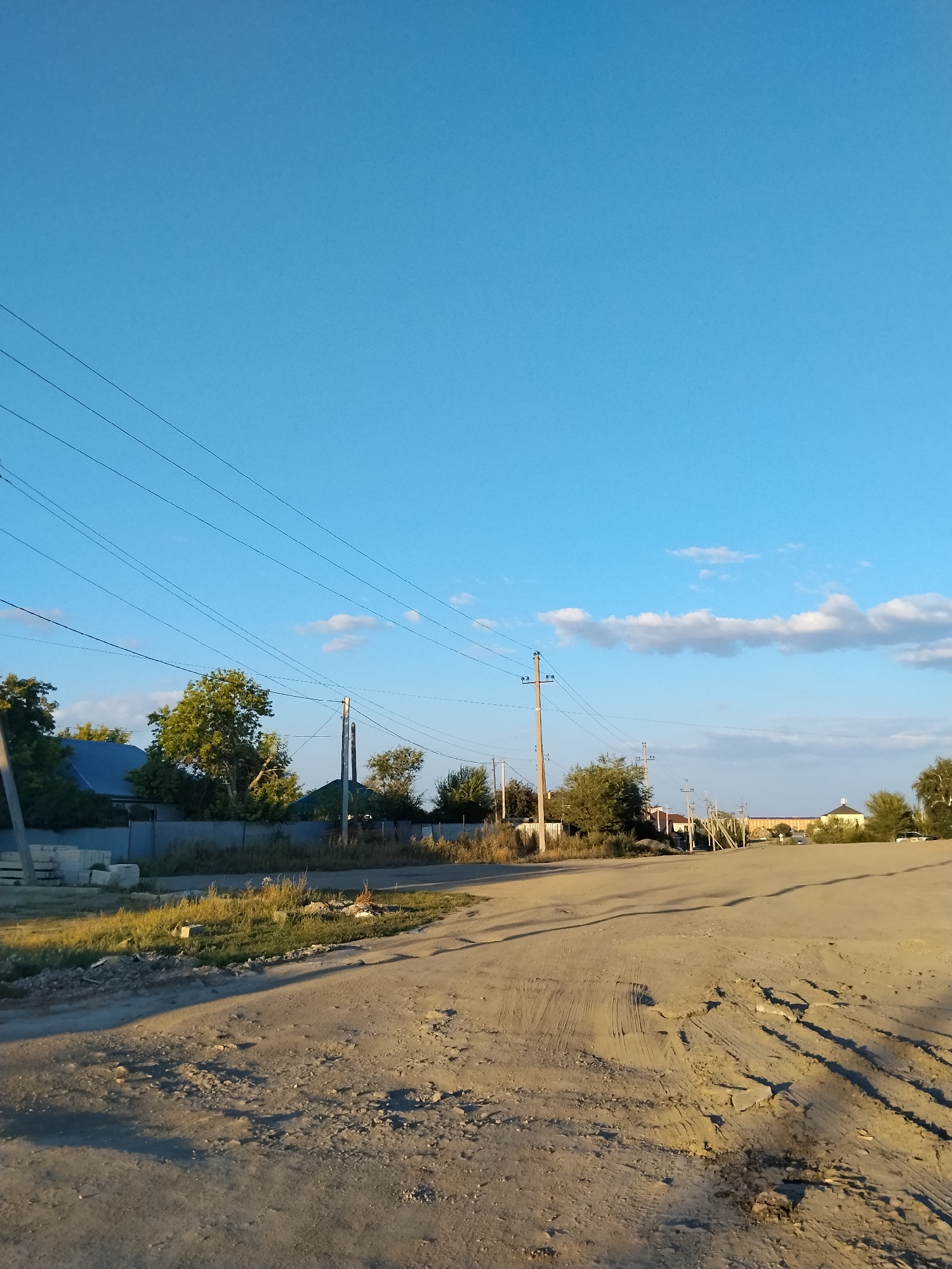 The image depicts a rural road scene in Kokshetau, Kazakhstan, under a clear blue sky with scattered white clouds. The road surface is unpaved and appears worn, with visible tire tracks and debris. Along the side of the road, there are power lines supported by wooden poles, typical of rural infrastructure. In the background, houses with blue and green roofs are visible, interspersed with trees. The overall impression is of a quiet, possibly arid, settlement during daylight hours.