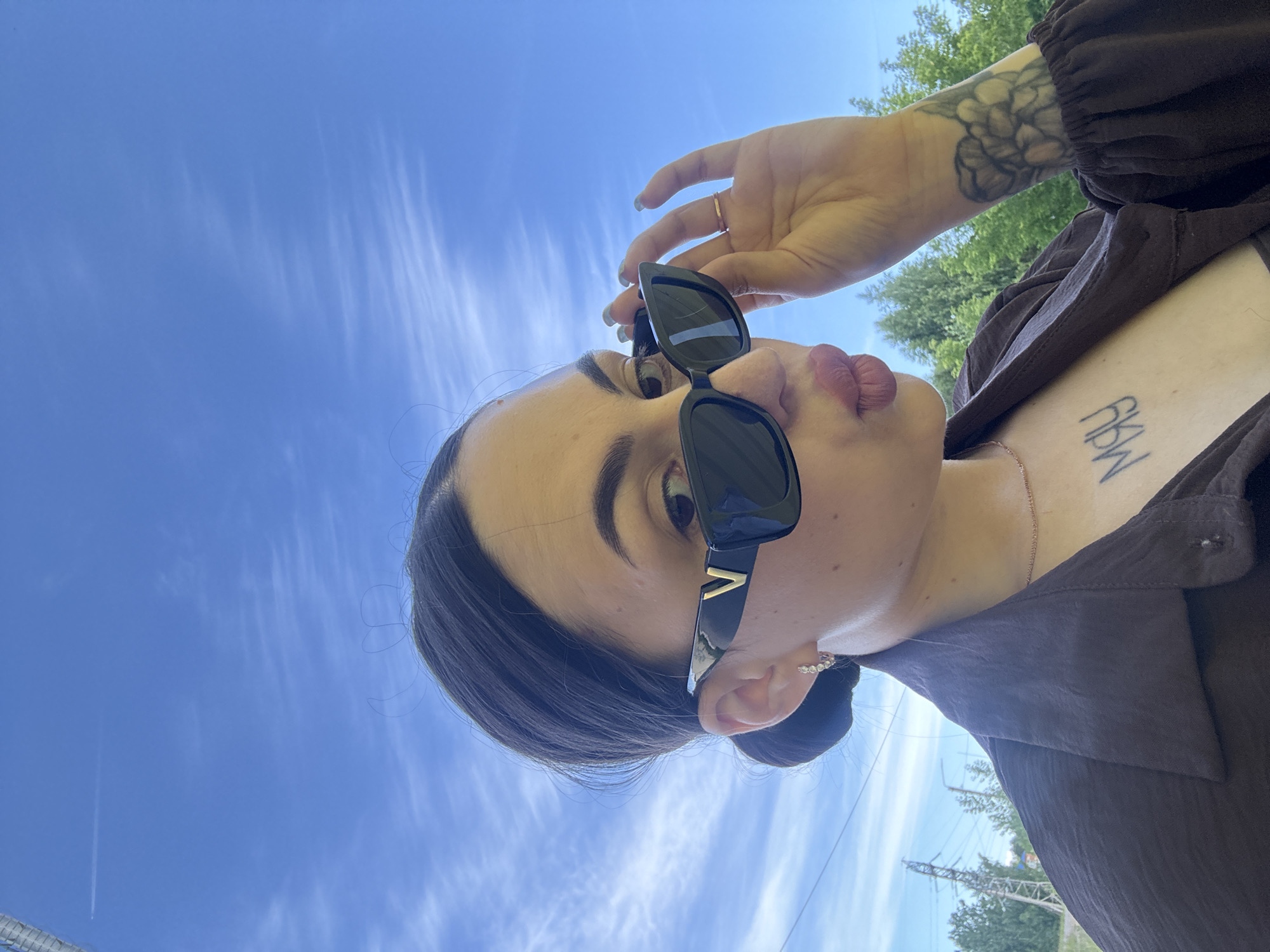 A young woman is taking a selfie outdoors against a bright blue sky with wispy clouds. She is wearing black sunglasses with a gold "V" logo on them and is adjusting them with her right hand. Her lips are pursed. She has dark hair pulled back into a bun and is wearing a brown collared shirt. A thin rose gold necklace is visible around her neck, and she has a tattoo of a flower on her right forearm and another tattoo on her chest that appears to say "MAY". In the background, there are power lines and lush green trees, suggesting an outdoor, possibly suburban or rural setting. The image does not provide specific landmarks to identify the exact location within Chaikovsky, Russia.