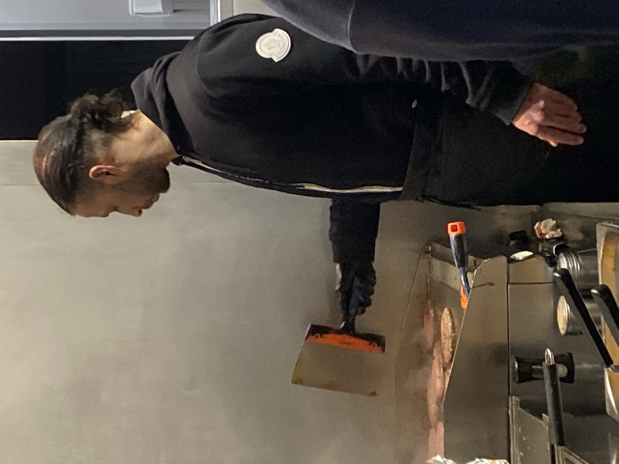 A person wearing a black jacket with a circular logo on the left shoulder is cooking on a flattop grill. The person is using a metal spatula with an orange handle to manipulate food on the grill. The location is Schiltigheim, France.