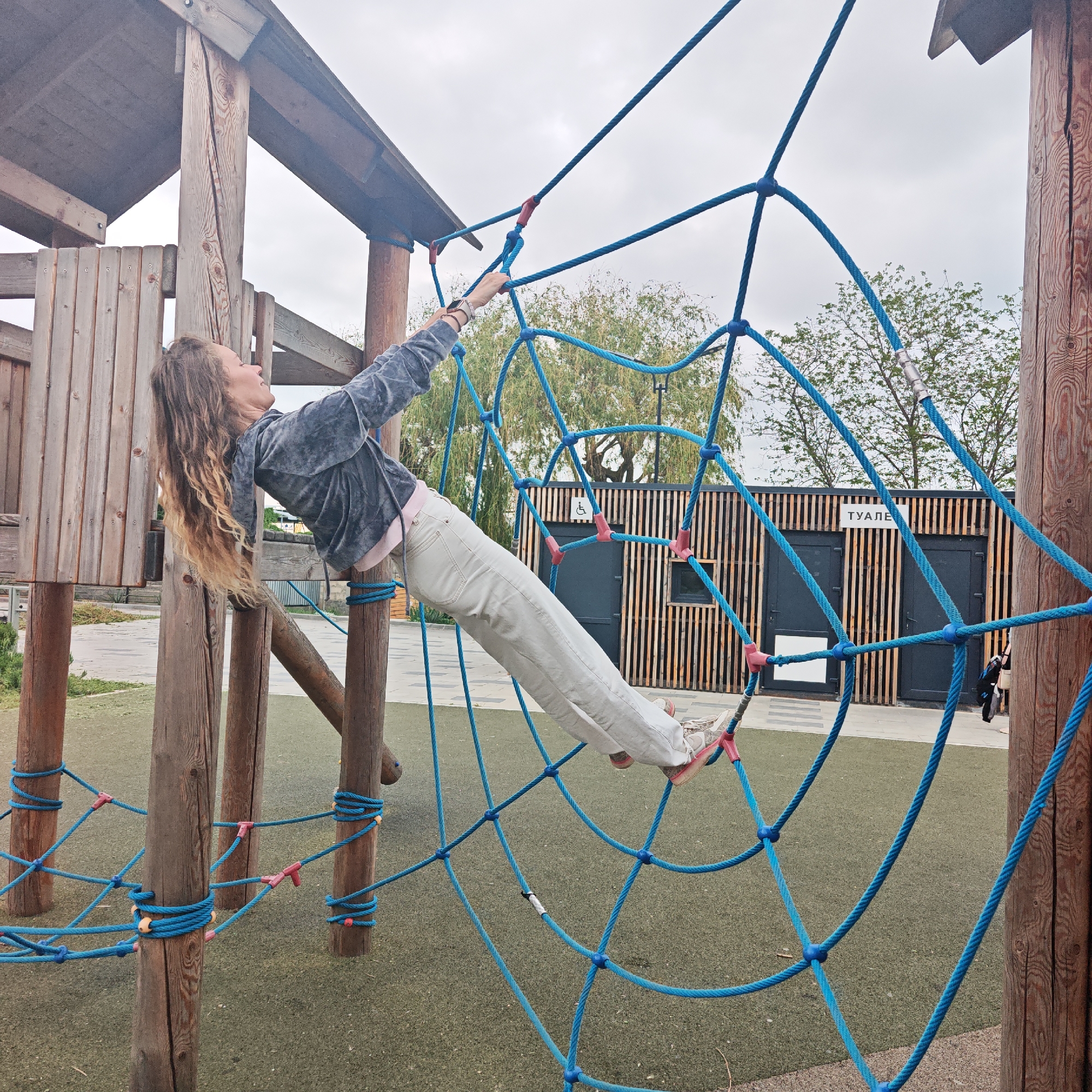 A woman is climbing on a blue rope climbing structure at a playground in городское поселение Щелкино, Ukraine. The structure is made of wooden posts and blue rope. The woman has long, curly, blonde hair and is wearing a gray velvet jacket, pink top, and white pants. Her left hand is above her head gripping the rope. Her right leg is extended with her foot supported by the ropes. There is a public toilet building nearby made of vertical wooden slats. The sky is overcast.