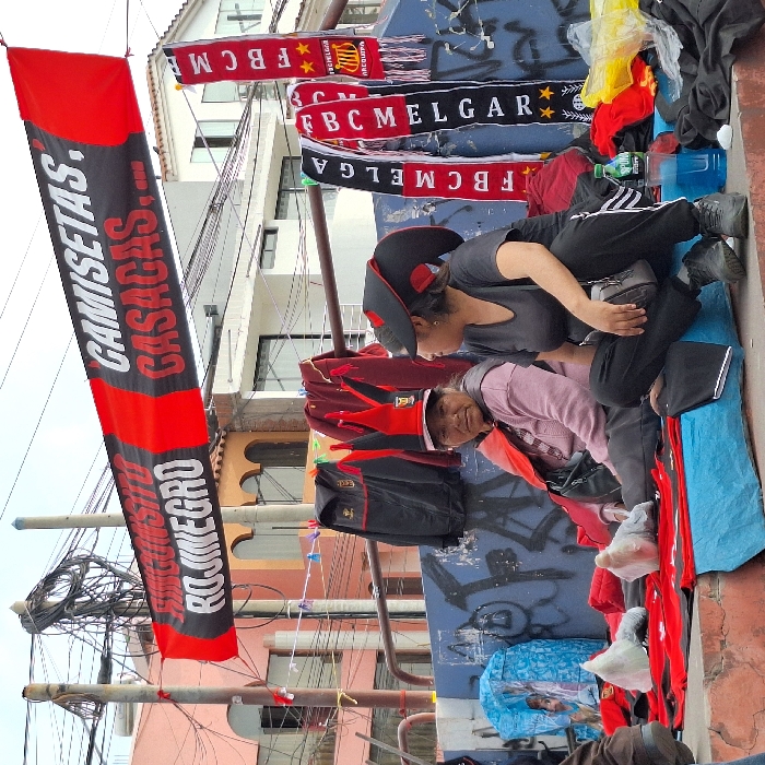 Two women wearing peaked hats are tending to merchandise spread out on a sidewalk. The goods include red and black clothing items, team scarves with “FBC MELGAR” printed on them, and plastic bags. A vertical banner advertises “CAMISETAS” and “CASACAS.” Electrical wires are visible overhead, and buildings form the backdrop to the street scene.