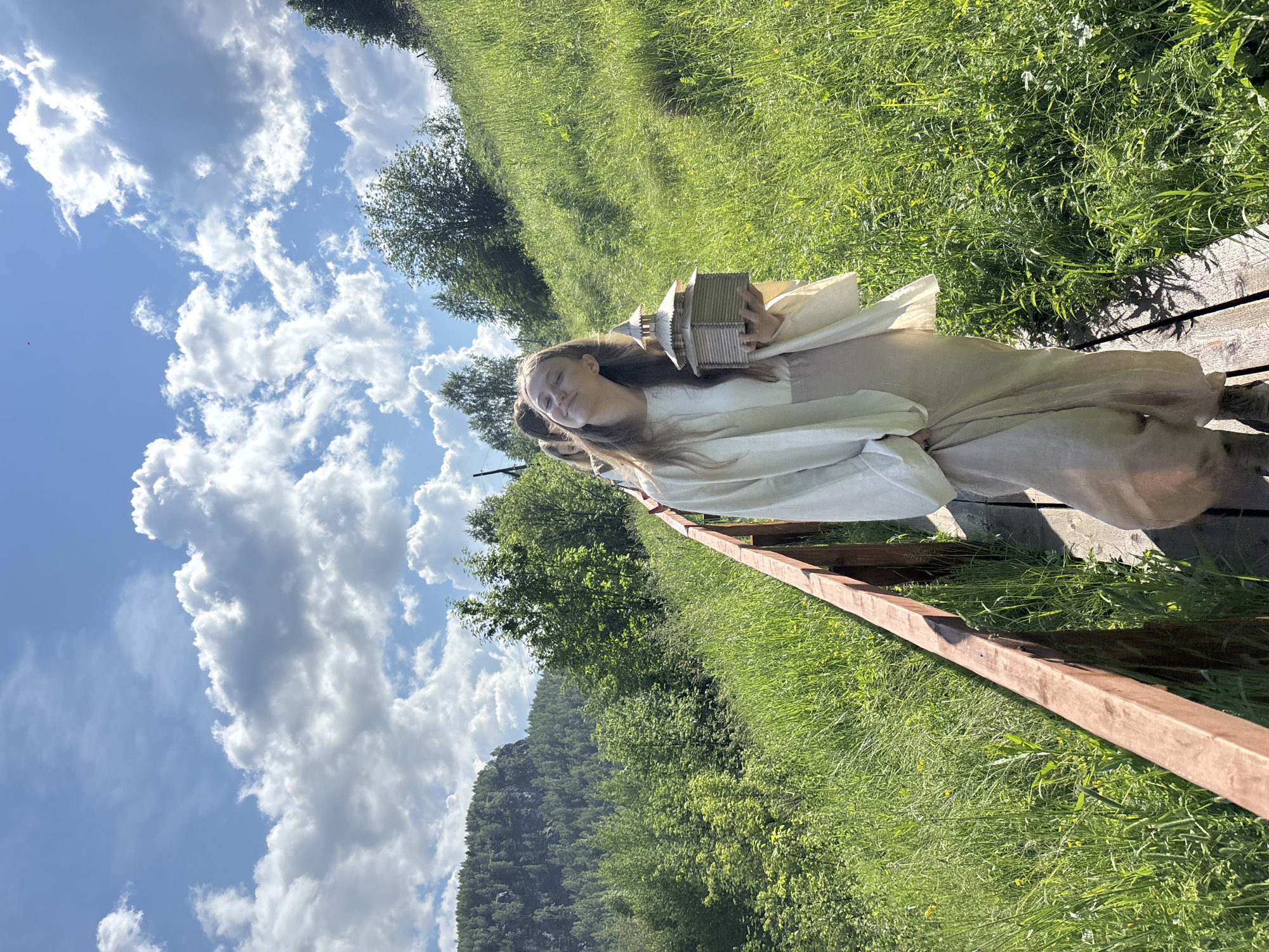 In Gubakha, Russia, a young woman with long brown hair walks along a wooden boardwalk. She is wearing a flowing cream-colored robe over a light-colored skirt and holds a miniature wooden building in her hands. She is smiling with her eyes closed, her head tilted back. Behind her, a few other people are visible on the boardwalk, walking in the same direction. The boardwalk is bordered by a wooden railing and surrounded by lush green grass and trees. In the background, a large, rocky mountain covered in trees can be seen under a bright blue sky with fluffy white clouds. The sun is shining, casting shadows on the boardwalk.