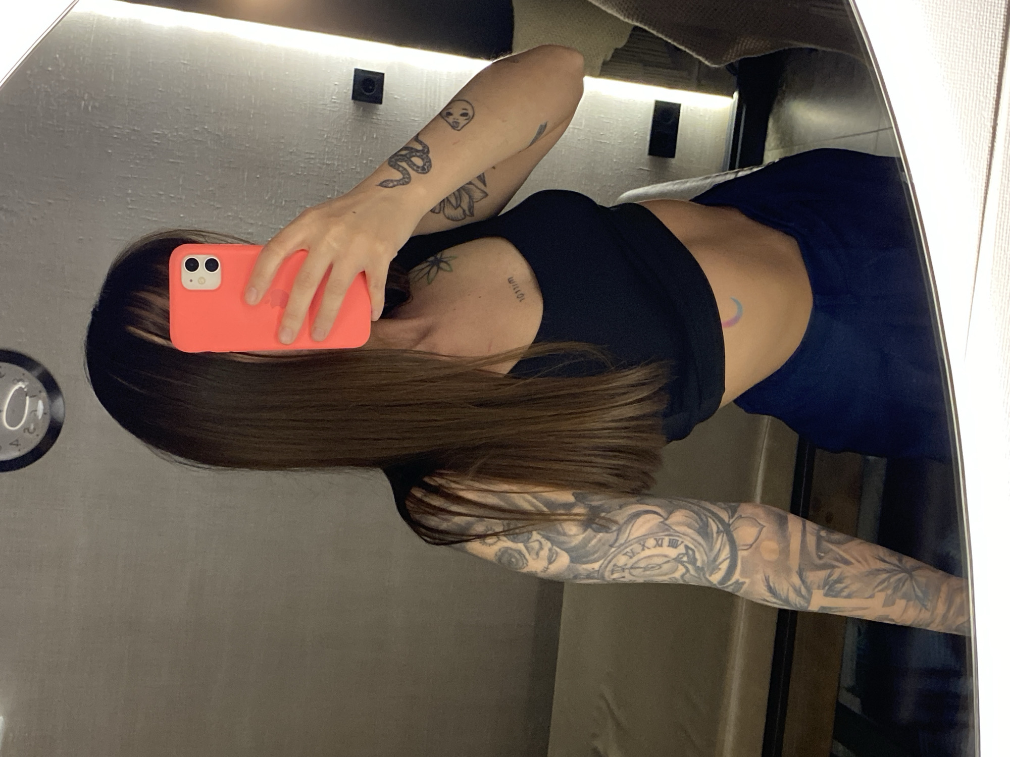 A person is captured in a mirror selfie, holding a bright orange smartphone case to cover part of their face. The image is oriented with the person's head to the left, appearing to be lying down or leaning. They have long, straight, dark brown hair.
The person is wearing a black crop top, revealing a midriff, and dark blue bottoms. Numerous tattoos are visible:
* On their right arm (viewer's left), there's a full sleeve featuring a detailed clock face with Roman numerals, floral patterns, and abstract designs.
* On their left arm (viewer's right), there's a snake, a small skull, a lotus flower, and other smaller designs.
* On the right collarbone area, a small flower tattoo and inverted text, possibly "WILLOW," are visible.
* On their left side/ribcage, a small, colorful crescent moon tattoo is present.
The background shows an indoor setting, likely a room in Votkinsk, Russia, reflected in the mirror. There are light-colored walls, possibly textured, with linear LED strip lighting illuminating the top edge of the wall. Dark, square fixtures, which appear to be wall outlets or switches, are mounted on the wall above the person's head. A wall-mounted clock with the numbers "0" and "1" visible is partially seen on the left side of the reflection. Part of a bed or sofa with a light-colored blanket is also reflected in the upper right. The overall impression is of a casual, personal moment captured indoors under artificial lighting.