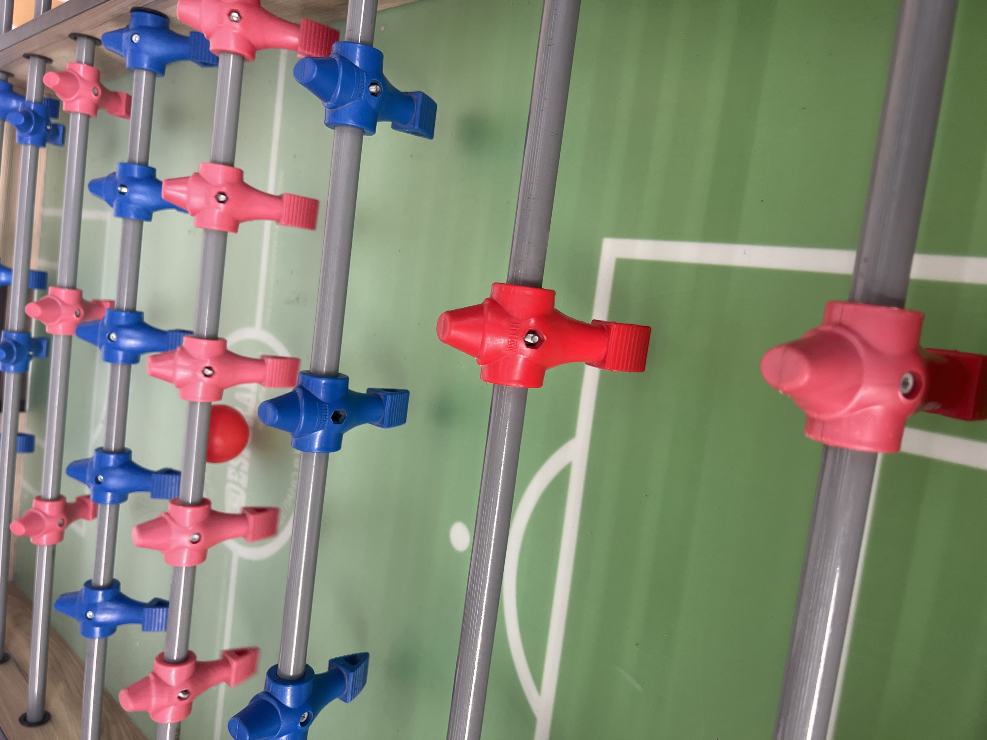 The scene captures a close-up, high-angle view of a foosball table's playing surface, located in Chaikovsky, Russia. The table features a green field with white markings, simulating a soccer pitch. Multiple horizontal gray metal rods traverse the playing area, supporting arrays of plastic player figurines. One team consists of blue players, one of which is marked "Made in Germany." The opposing team comprises pink/red players, with at least one bearing the inscription "DESPERADO." A single red foosball ball rests centrally on the green field, positioned between a blue player and a pink/red player. Portions of a light brown, possibly wooden, table frame are visible at the upper and right edges. The playing surface exhibits minor debris or wear. No human presence or active play is depicted.