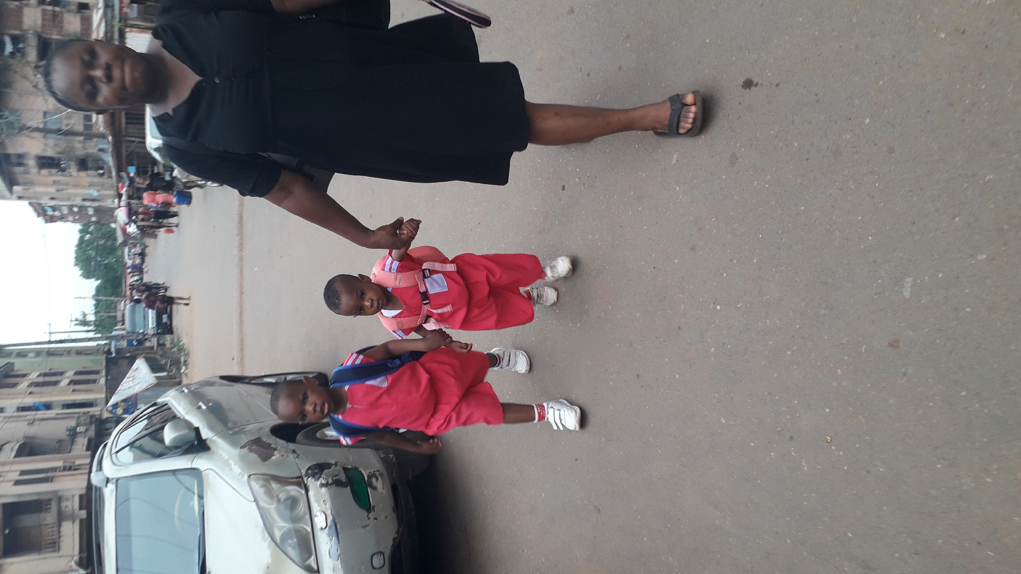 The image shows a woman walking on a street in Onitsha, Nigeria, holding the hands of two young children wearing matching red uniforms, white socks, and white sneakers. The children also have backpacks. The woman is wearing a black dress and sandals. A silver car with visible damage is parked on the side of the street. The background includes buildings and other people, suggesting a bustling urban environment.