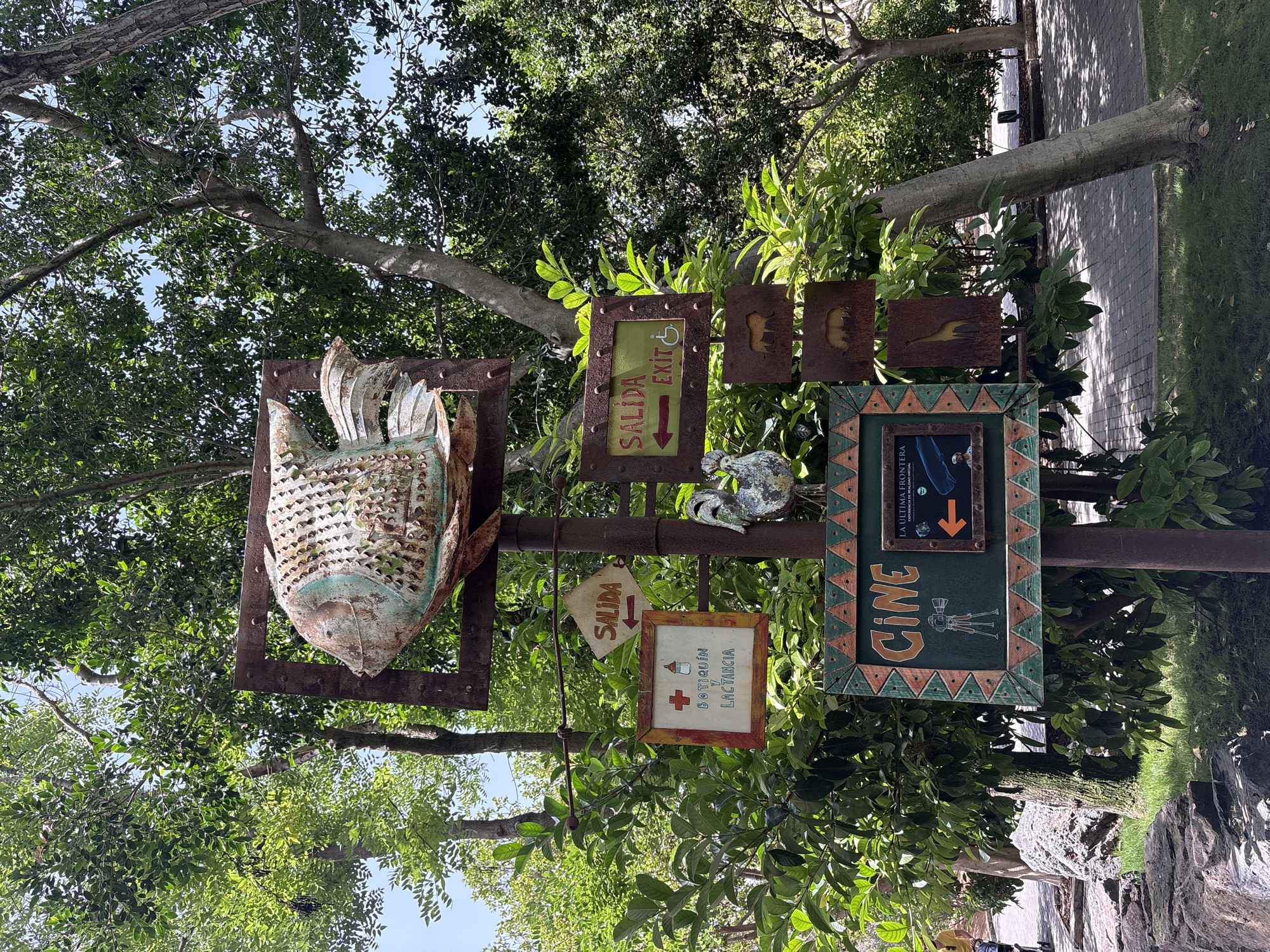 This image depicts a whimsical signpost in Valencia, Spain, likely within a park or attraction. The signpost is adorned with a large, stylized metal fish sculpture at the top, weathered and showing signs of age. Below the fish, several signs are attached to a central pole. One sign, a diamond shape with a red border, points to the left and reads "SALIDA" (Exit). Another sign, rectangular with a green background and red border, has a white cross and a baby bottle symbol, indicating "BOTIQUÍN Y LACTANCIA" (First Aid and Nursing). To the right, a rectangular sign with a green background reads "SALIDA EXIT" with an accessibility symbol. Further down, smaller, rusted metal plates are attached, featuring silhouettes of animals like a horse and elephants. At the bottom, a large, dark green sign with a decorative border displays "CINE" in orange lettering, accompanied by a graphic of a vintage film camera on a tripod. To the right of "CINE," a smaller framed sign shows an orange arrow pointing left and text that appears to be "LA ÚLTIMA FRONTERA EXPERIENCIA DE REALIDAD VIRTUAL" (The Last Frontier Virtual Reality Experience). The signpost is surrounded by lush green trees and foliage, with dappled sunlight filtering through the leaves. A paved walkway and grassy areas are visible in the background, suggesting an outdoor public space. The overall atmosphere is one of a well-maintained, family-friendly attraction.