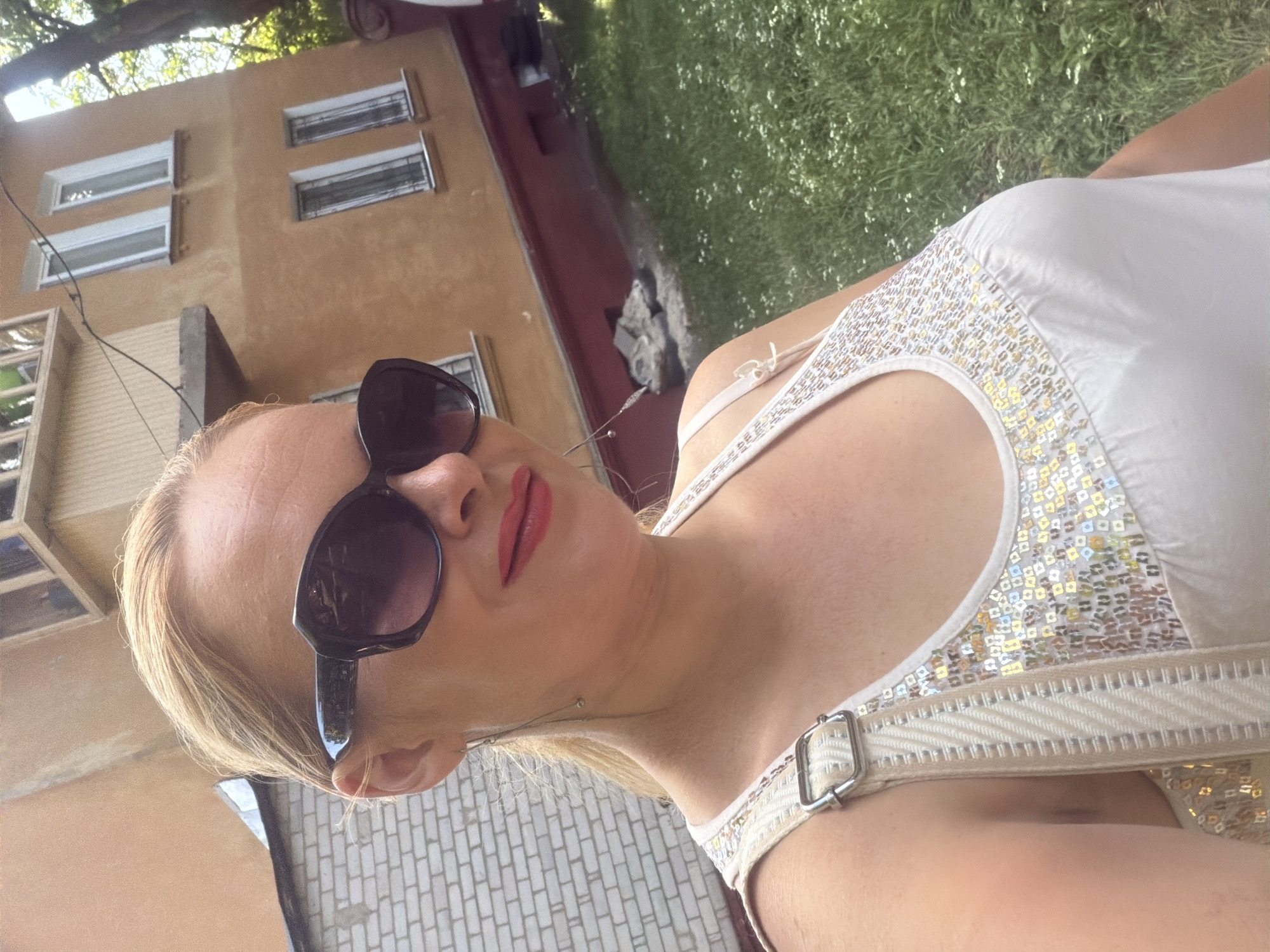 A blonde woman, wearing a white tank top adorned with sequins and large sunglasses, poses for a selfie outdoors. She has bright red lipstick on. In the background, an orange building with white-trimmed windows stands next to a white brick wall. Green grass with scattered white flowers covers the ground in front of the building. The scene appears to be an urban residential area.