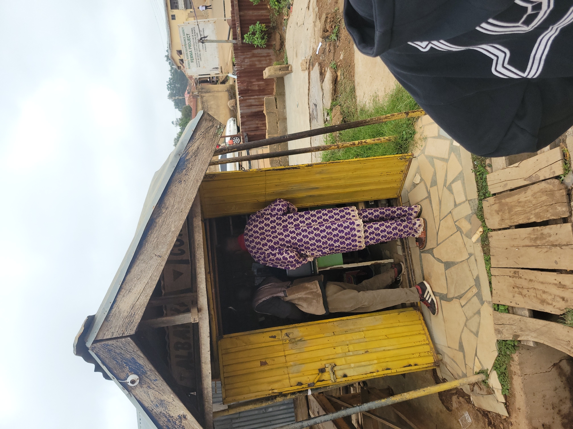 The image depicts a street-level view in Jos, Nigeria, featuring a small, open-fronted shop with a yellow metal door and a rustic wooden structure above. Two individuals are visible inside and at the entrance of the shop. One person, wearing a purple and white patterned outfit, stands facing away from the camera, partially inside the shop. Another person, wearing a dark vest over a shirt and light-colored trousers, is also within the shop, facing away from the camera. The shop is situated on a paved area made of irregular stone tiles. In the background, a white sign with text is visible, identifying it as the "FEDERAL ROADS MAINTENANCE AGENCY FERMA PROJECT." The surrounding environment appears to be a dusty, unpaved area with some vegetation and partially constructed brickwork. A portion of a person wearing a black hoodie with a white graphic design is in the foreground, on the right side of the image, obscuring part of the scene. Wooden planks are laid out in the lower foreground, suggesting a makeshift walkway over a ditch or uneven ground. The sky is overcast.