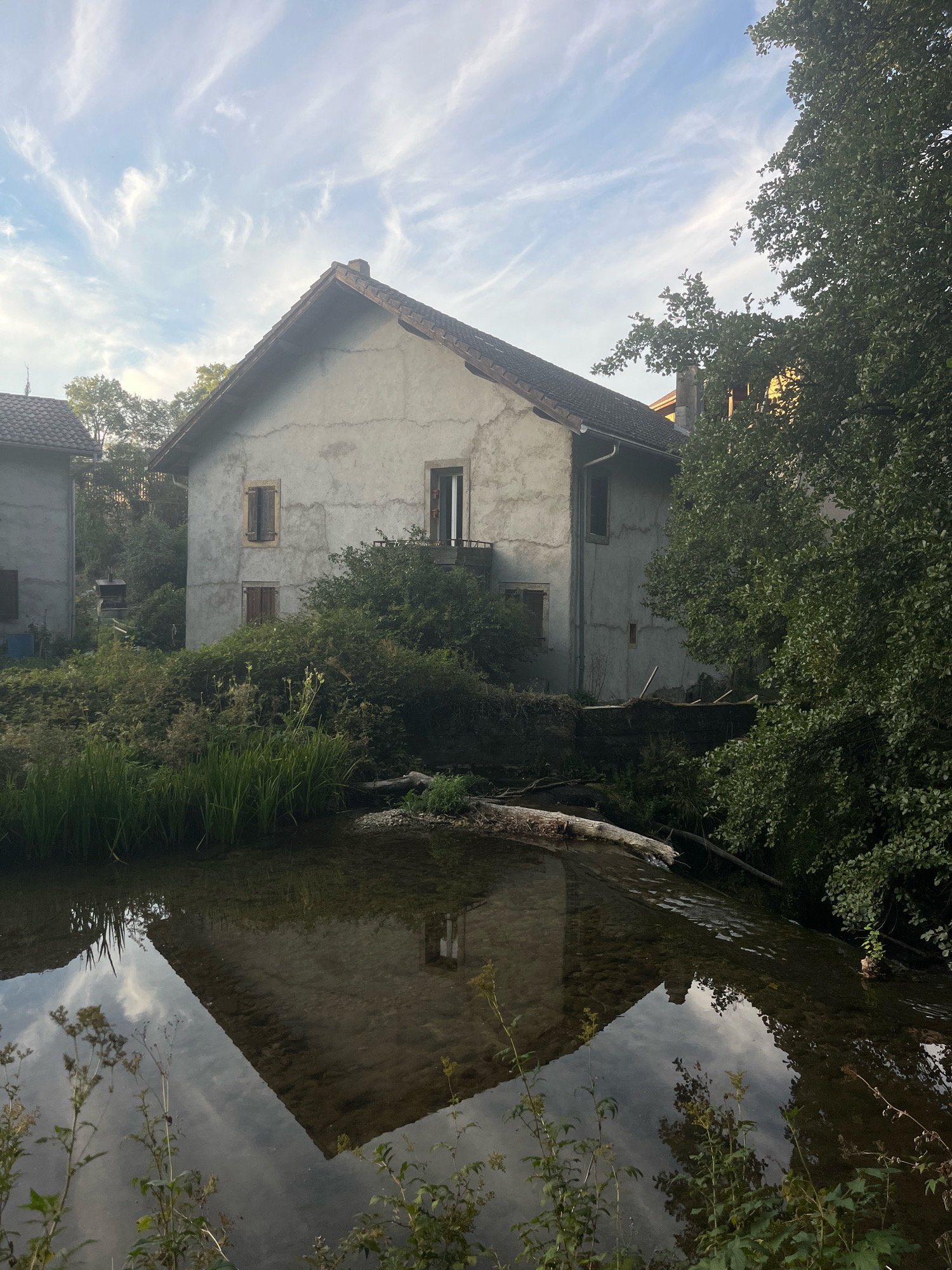 This image depicts a rustic, light-colored building with a pitched roof, situated in the countryside of Chevilly, Switzerland. The building appears to be two stories high with several windows, some of which have wooden shutters. It is surrounded by dense green foliage and trees, suggesting a natural, possibly overgrown environment. In the foreground, a body of water, likely a stream or river, reflects the sky and the building. The water is shallow, revealing a pebble-strewn bottom and a fallen log. The sky is partly cloudy with wispy white clouds set against a light blue background, indicative of daytime. No people are present in the scene. The overall atmosphere is tranquil and serene, capturing a moment of rural charm.