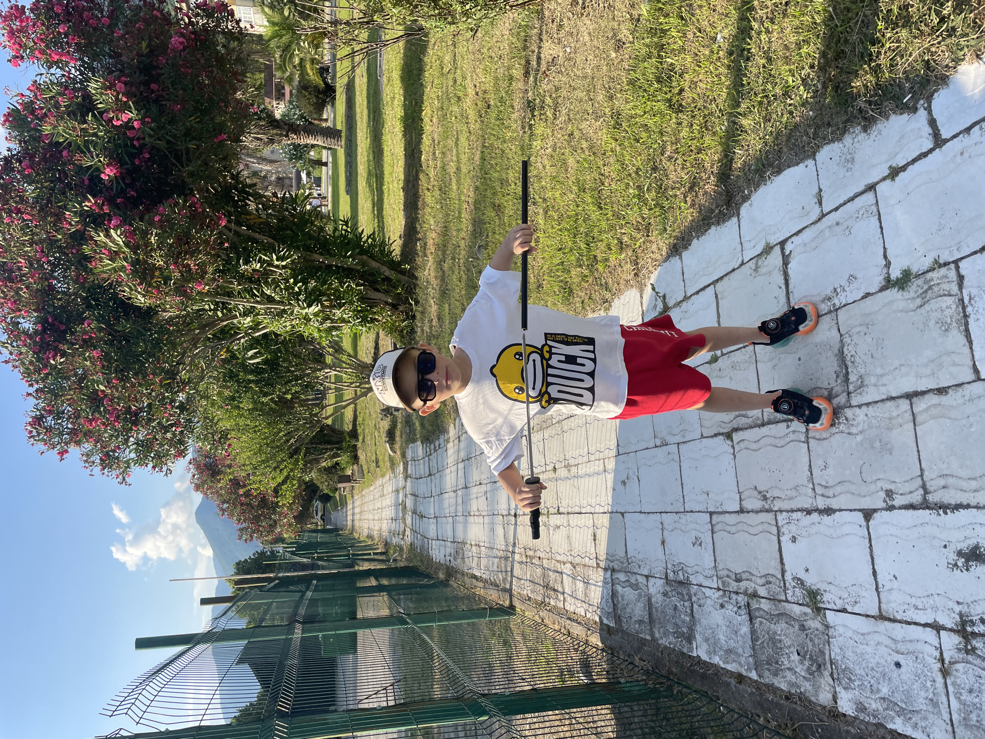 A young boy stands on a paved walkway in Gagra, Abkhazia, holding a dark rod horizontally. He is wearing a white t-shirt with a yellow duck cartoon and the word "duck" printed on it, red shorts, sunglasses, a beige baseball cap, and black and orange athletic shoes. The walkway is lined with a green metal fence on the left and a grassy area with trees and pink blooming bushes on the right. In the background, mountains are visible under a clear blue sky with scattered white clouds. The scene appears to be a park or a public area with lush vegetation and paved paths.