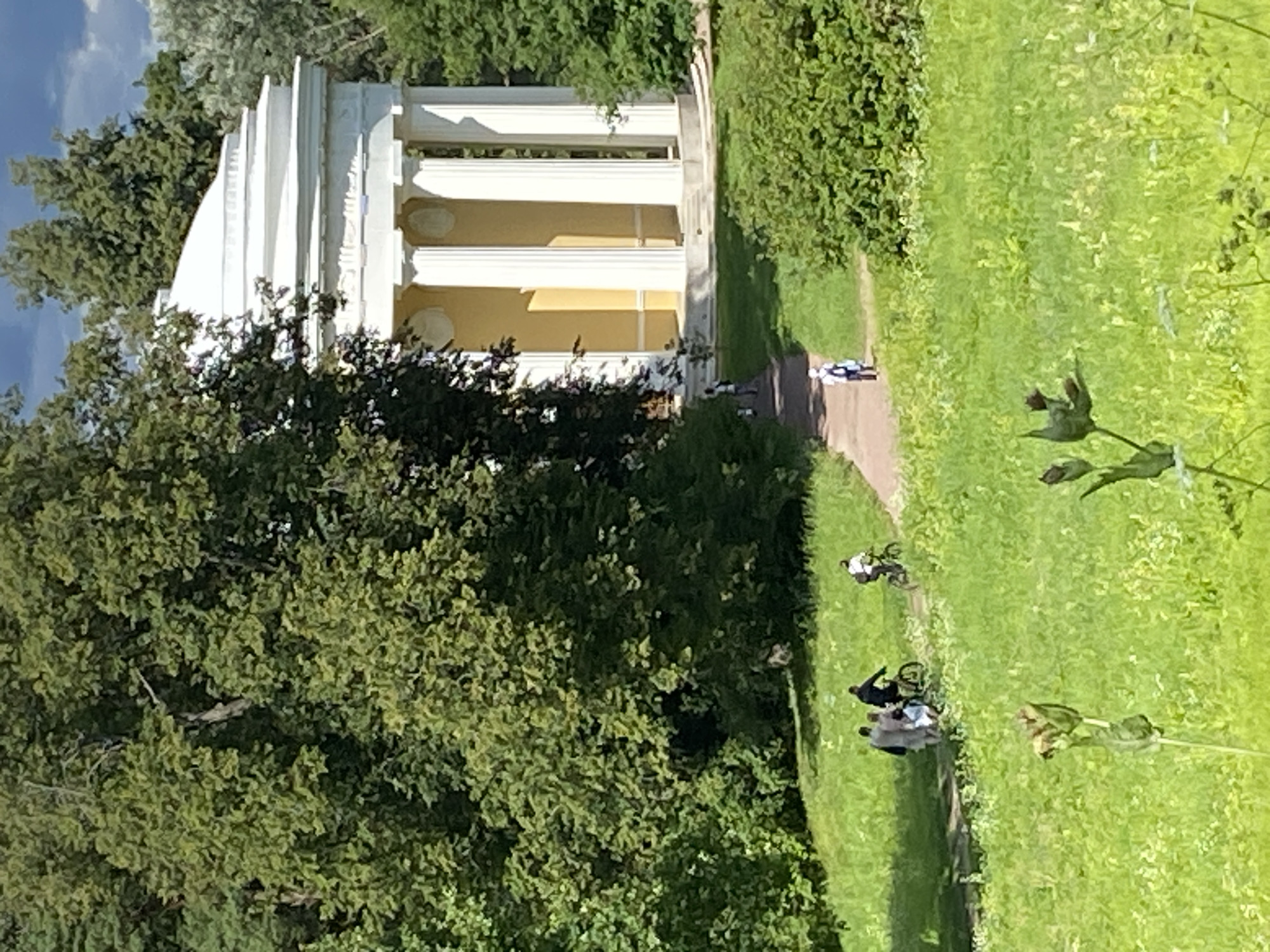 The image depicts a sunny day in the Pavlovsk Park in Pavlovsk, Russia. In the background, a bright yellow building with white classical columns and a white domed roof is visible, surrounded by lush green trees. In the mid-ground, a paved path winds through a grassy area. Several people are seen on the path. On the left, a group of three individuals, two walking and one on a bicycle, are moving away from the camera. Further up the path, another person is on a bicycle, also moving away. In the foreground, the scene is dominated by a vibrant green lawn with scattered wildflowers and some plants with seed pods. The overall impression is of a tranquil park scene with people enjoying outdoor activities.