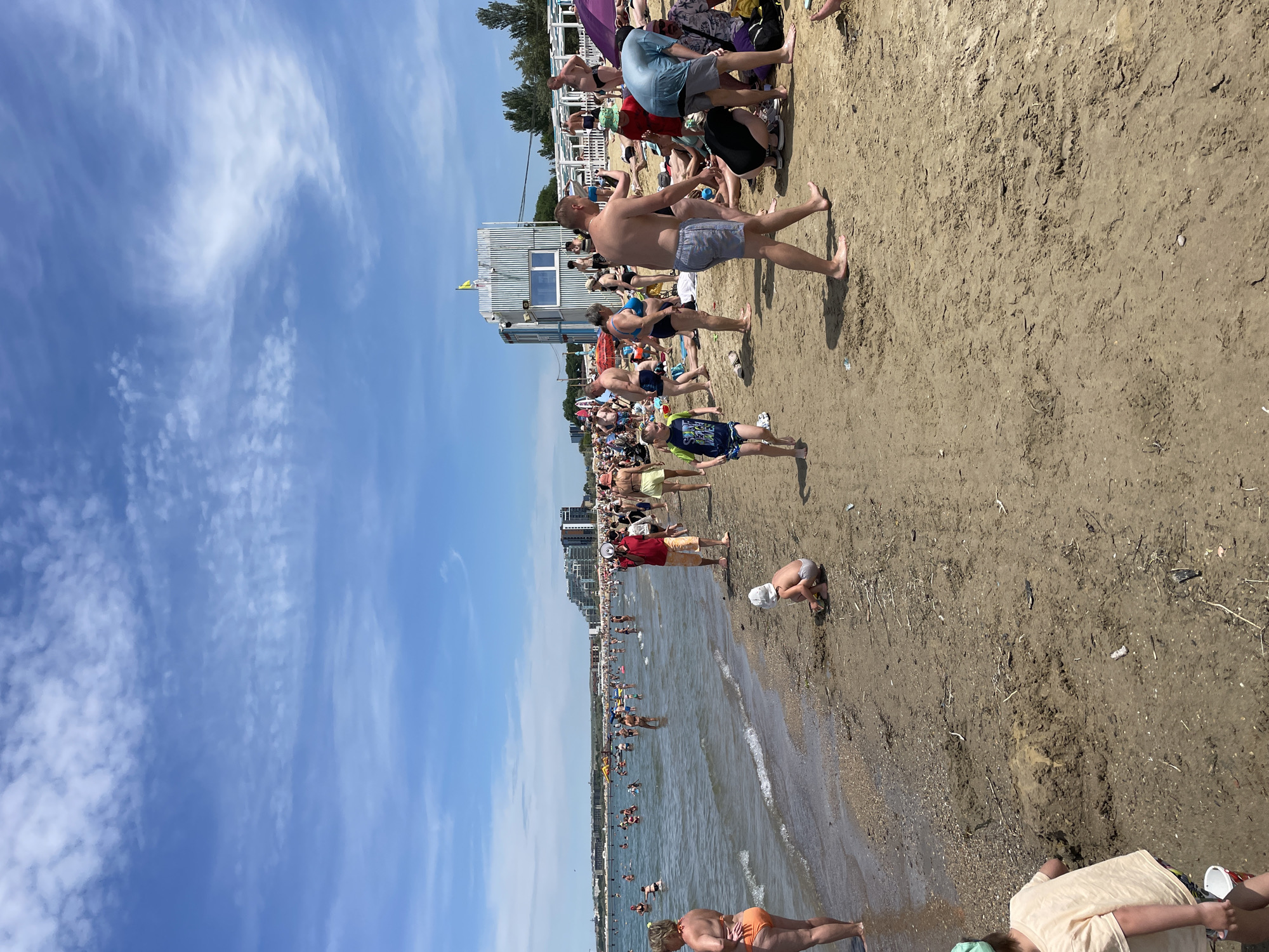 The image depicts a crowded beach scene under a bright blue sky with scattered white clouds. The shoreline is populated with numerous people engaging in various activities. In the foreground, the sandy beach is visible with some debris. A young child in a light-colored t-shirt and a hat is playing in the sand. Further back, many adults and children are in the shallow water, wading, swimming, and playing. A lifeguard stand with a green and white striped structure and a yellow flag is visible on the beach. In the background, there are more people sunbathing and walking on the sand, as well as some buildings and trees along the coastline. The overall scene conveys a busy summer day at a popular beach resort.