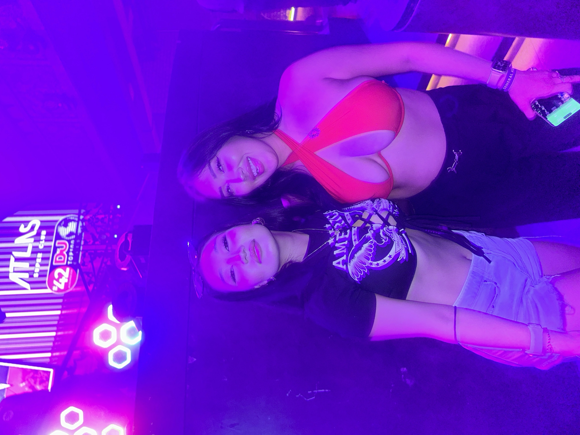 Two women are standing in a nightclub illuminated by purple and pink lights. The woman on the left is wearing a black t-shirt with a graphic design and light-wash denim shorts. She has long dark hair and is looking towards the camera with a neutral expression. She is wearing sunglasses on her head and a necklace. The woman on the right is wearing a bright red halter-neck bikini top and black pants. She has long dark hair and is smiling at the camera, with her tongue sticking out slightly. She is wearing a watch and bracelets. In the background, a neon sign reads "ATLAS SUPER CLUB". The overall environment appears to be a lively nightclub setting.