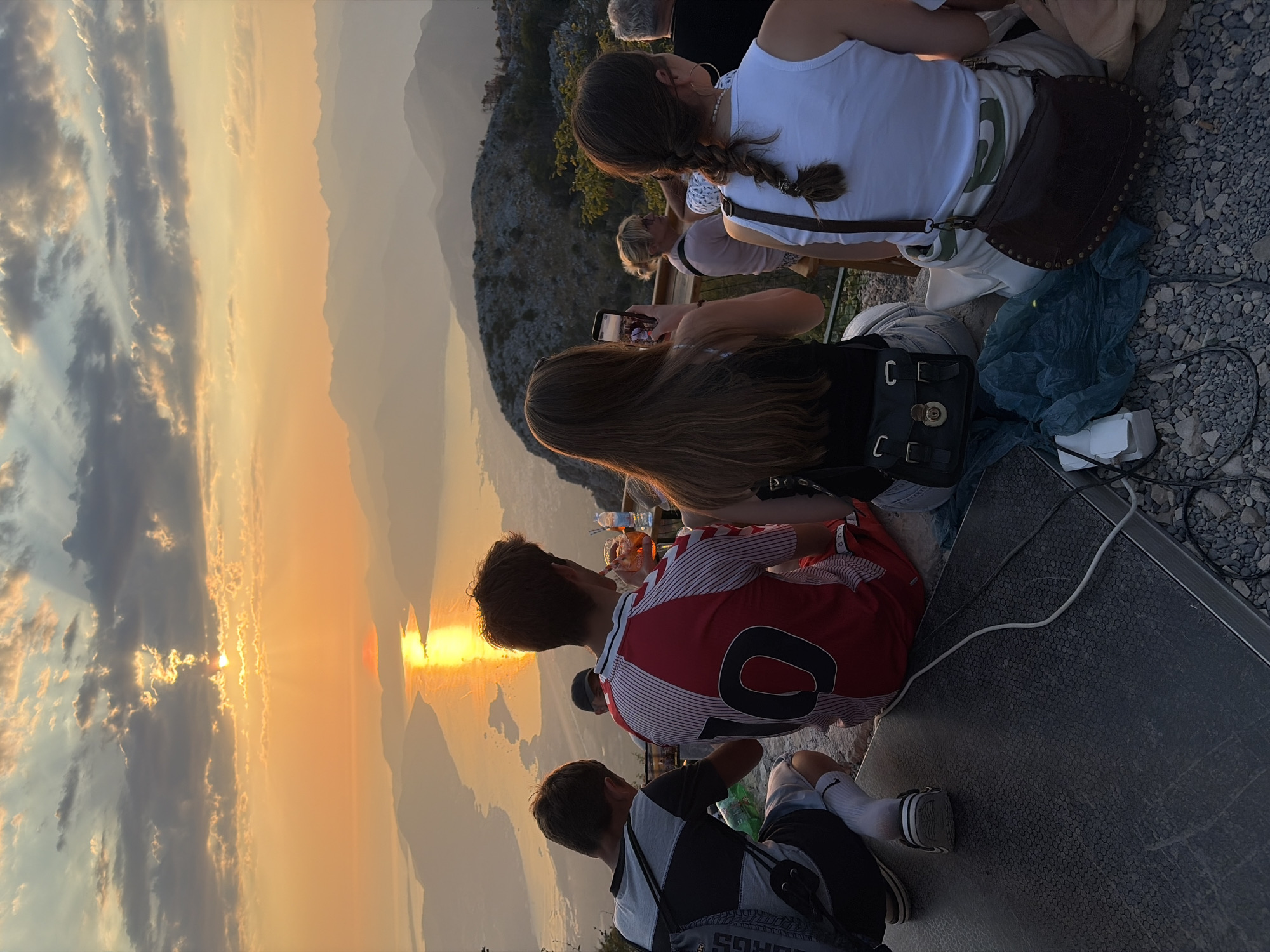 The image depicts a group of individuals seated on a stone ledge overlooking a scenic landscape at sunset. The sky is ablaze with warm hues of orange and yellow, with sunlight streaming through dramatic clouds, illuminating a body of water and distant mountains. In the foreground, four people are visible from behind. The person on the far left is a young male wearing a grey and black striped t-shirt, white shorts, and dark sneakers. He has a backpack slung over his shoulder. Next to him is another young male in a red and white athletic jersey with the number "10" on the back, holding a drink. To his right, a young female with long brown hair is holding up a smartphone to take a picture. Further to the right, another female with braided brown hair is wearing a white tank top. The background reveals a vast expanse of layered mountains fading into the hazy distance, with the glowing sun reflected on the water below. The setting appears to be an elevated viewpoint, possibly a tourist spot, given the presence of a wooden railing and the general ambiance of people enjoying a view. The location is identified as Kuk, Montenegro, suggesting a mountainous coastal region.