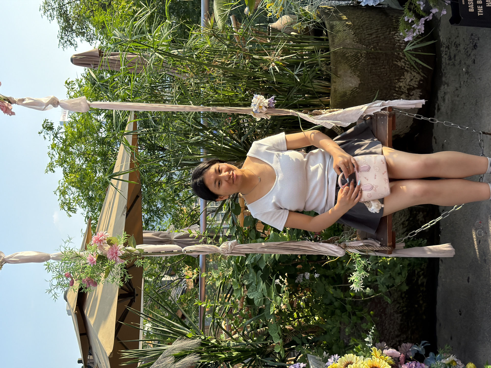 A young Asian woman with dark hair is sitting on a wooden swing in an outdoor setting, likely a cafe or garden area, in Yuzhong District, China. She is wearing a white t-shirt and a dark skirt, holding a small pink handbag in her lap. The swing is adorned with fabric and artificial flowers. Behind her, there is lush greenery, including tall plants and trees, as well as an umbrella and some decorative elements. The lighting suggests it is late afternoon or early evening, with a clear blue sky visible. The ground appears to be a paved surface.