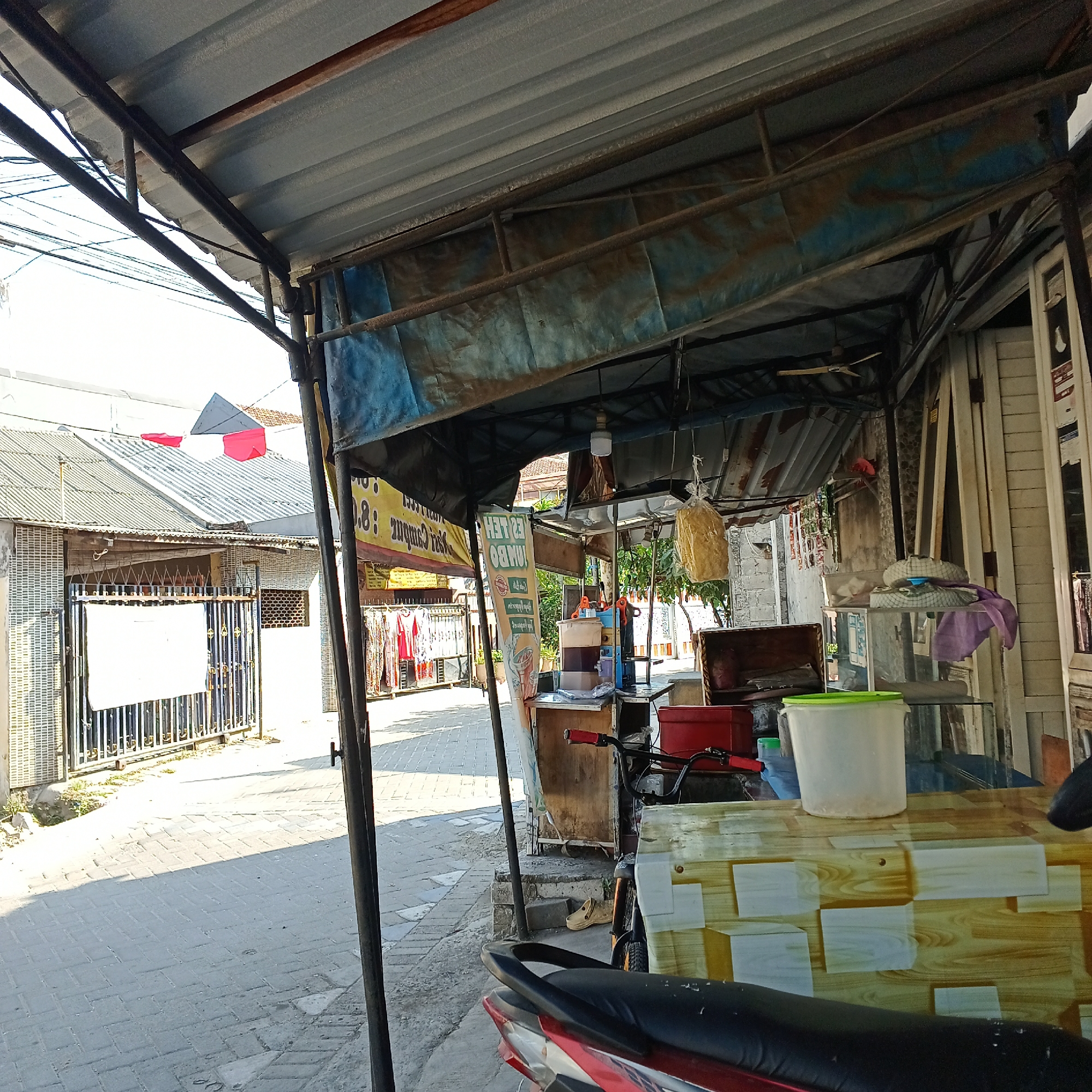 The image depicts a street-level view of a food stall or small shop in Kepuh Kiriman, Indonesia. The stall is covered by a corrugated metal roof supported by dark metal poles. A blue tarp is visible beneath the roof. In the foreground, the seat of a red and black motorcycle is prominent. To the left of the motorcycle, the pavement is paved with light-colored interlocking bricks, with a shadow cast across it. Beyond the pavement, a metal gate with vertical bars leads to a building with a tiled roof. Red and white triangular flags are attached to wires above the building. The stall itself has a wooden counter with a laminated surface featuring a wood-grain pattern. On the counter are various items including a large white plastic bucket with a green lid, a red cooler, and what appears to be a beverage dispenser. A bicycle is parked near the stall. Behind the stall, more buildings are visible, along with some greenery and hanging items that might be food products. A vertical banner with text in a foreign language stands near the stall. The overall scene suggests a commercial area on a sunny day.