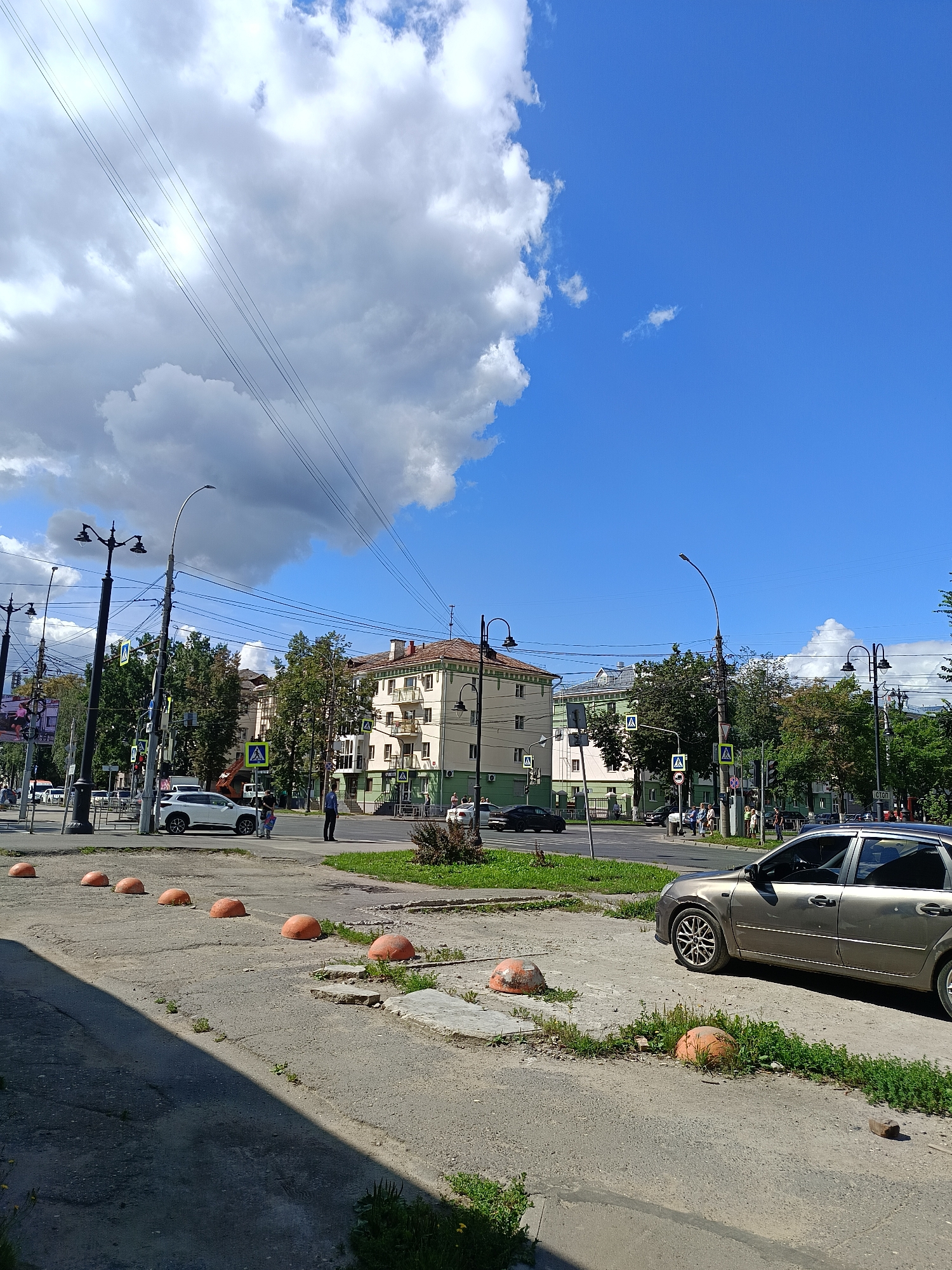 A street corner scene unfolds under a bright, blue sky with scattered white and dark clouds. In the foreground, a light-colored car is parked on a cracked asphalt surface, with several orange, spherical bollards lining the edge of the road. A row of similar bollards extends into the background. Beyond the bollards, a grassy area separates the street from a sidewalk where pedestrians are visible. A white SUV is stopped at what appears to be an intersection, with a man and a woman standing nearby. Across the street, several multi-story buildings are visible, some with balconies. Trees line the street, and streetlights are positioned along the sidewalks. Power lines crisscross the sky above the scene. The overall impression is a typical urban streetscape on a sunny day.