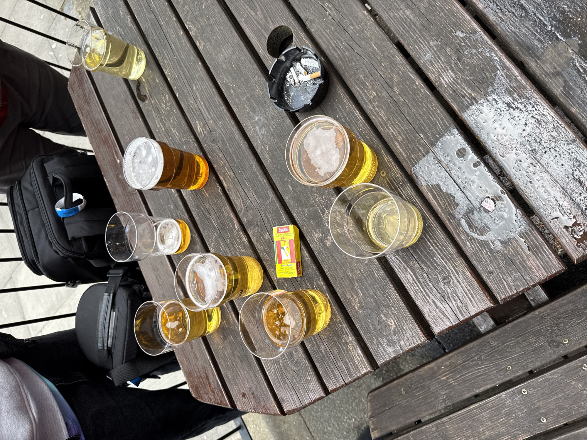 A high-angle shot shows a wooden picnic table, likely outdoors, littered with nine glasses filled with varying amounts of light-colored beverages, possibly beer or cider. Beside the glasses are an ashtray containing cigarette butts and a yellow box of Swan lighter fluid. A black bag and a glimpse of people's clothing are visible on the left side of the frame. The table is wet in places, suggesting recent rain or condensation. The location is implied to be in London, United Kingdom.