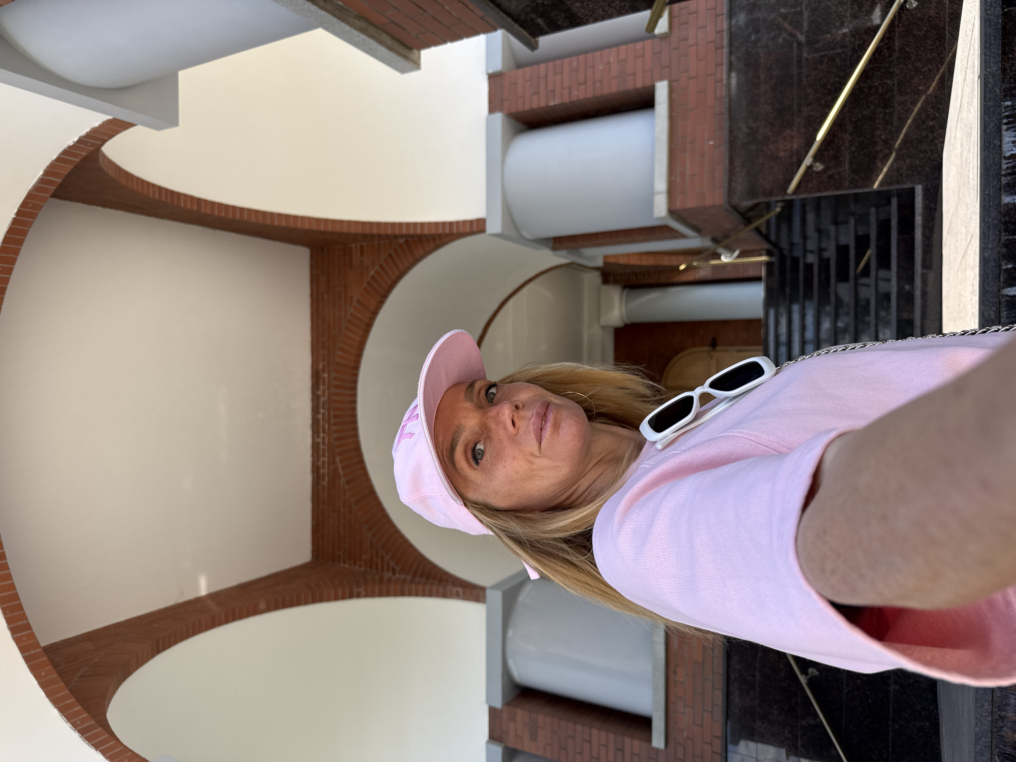 The image features a woman posing for a selfie in what appears to be an outdoor, architectural setting. She is wearing a pink baseball cap, a pink sweatshirt, and white sunglasses perched on her head. Her blonde hair is visible. The background consists of curved brick archways and light-colored walls, with some darker, modern architectural elements and a glimpse of metal railings in the distance.
The lighting suggests it is daytime, with sunlight casting shadows. The overall mood conveyed by the woman's expression and attire seems casual and perhaps playful. The location cues, such as the distinct architectural style with brickwork and rounded openings, combined with the provided context of Saint Petersburg, Russia, hint at a specific type of building or public space within the city. There is no visible text in the image.