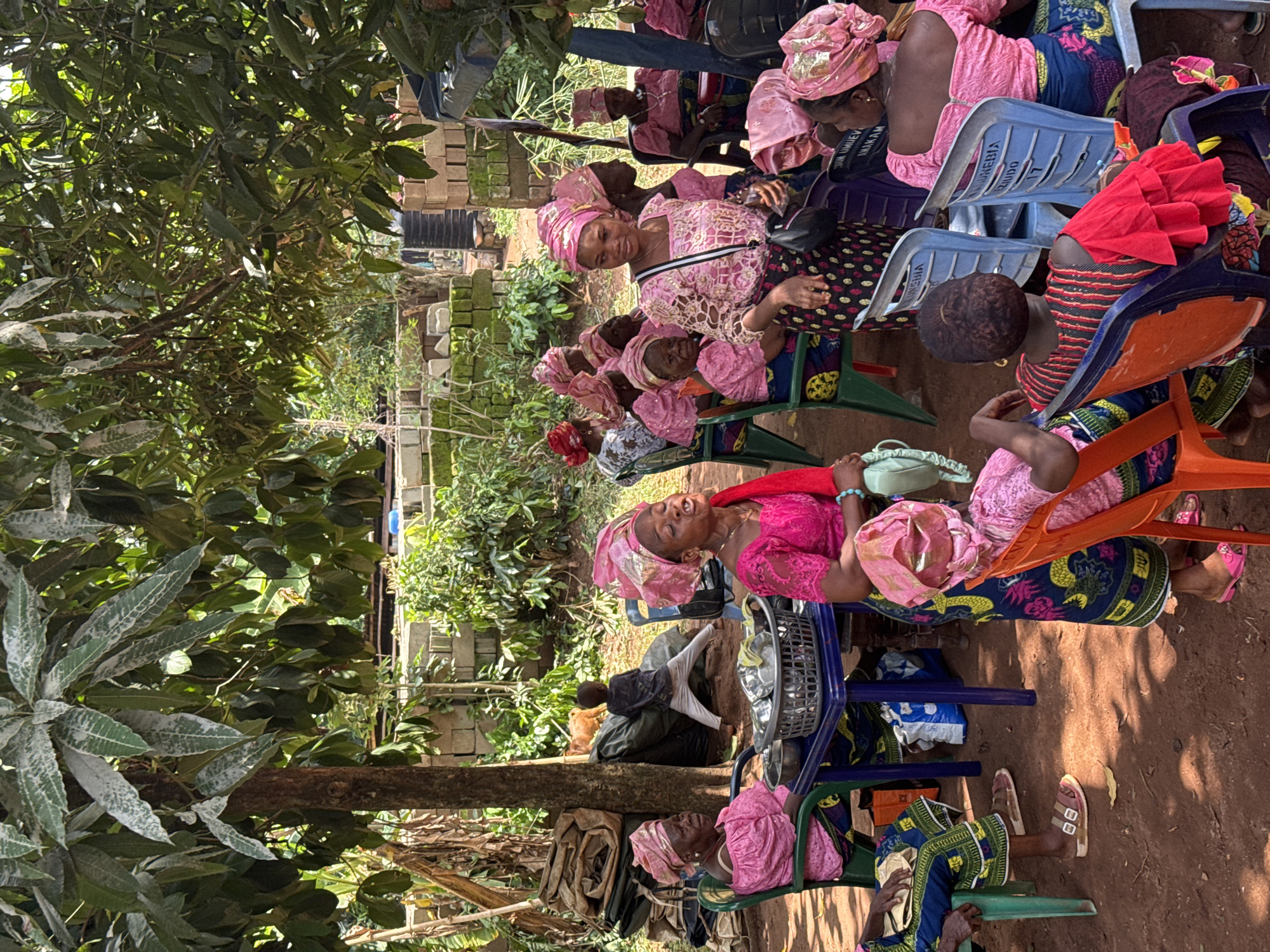 This image captures an outdoor gathering of women in Eziudo, Nigeria. The scene is set under the shade of lush green trees, with the ground composed of reddish-brown dirt. Several women, predominantly adults, are seated on an assortment of brightly colored plastic chairs (blue, green, and orange), arranged in an informal group.
Most of the women are dressed in vibrant traditional attire, featuring patterned fabrics for their skirts and blouses, often complemented by matching head wraps. Pink is a prominent color in many of their outfits and head wraps. One woman, centrally positioned and wearing a pink lace top with a matching head wrap, looks directly at the camera with a smile. Others are engaged in conversation or observing their surroundings. A child's head is visible among the seated adults, and another child is partially seen in the background on the far right.
A metal basket or tray containing various items rests on the ground near the center of the gathering. In the background, simple brick or block structures are visible amidst more foliage. The bright daylight suggests it is midday or early afternoon.
Visible text provides location cues: the word "EZIUDO" and "NNEBIA" along with the number "17" are printed on some of the blue plastic chairs. The word "MMANAM" is also faintly visible on a headscarf worn by one of the women. The overall ambiance is that of a social and calm community event.