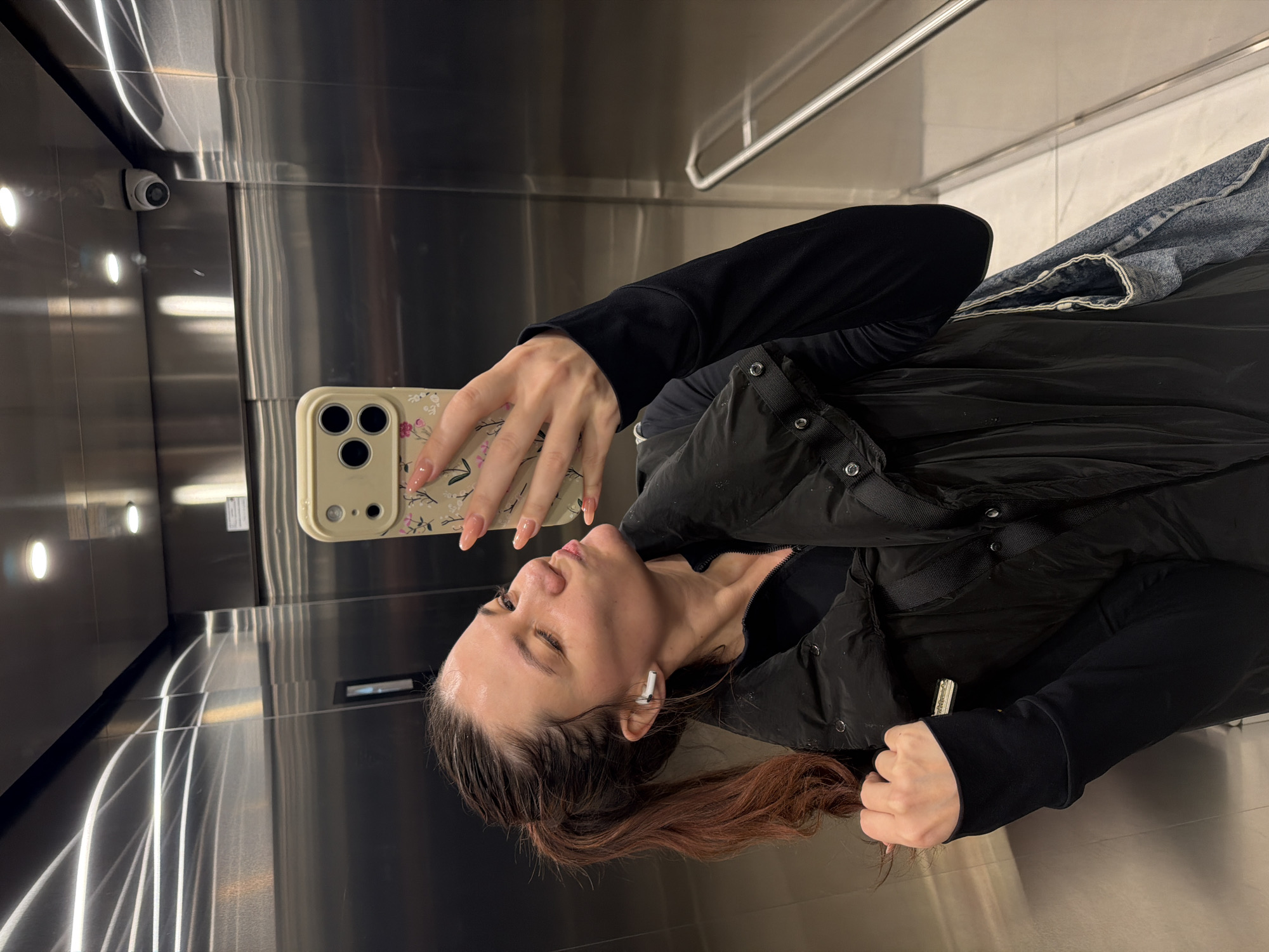 A woman is taking a selfie in an elevator. She is holding a smartphone in her right hand, and the phone's case has a floral design. The woman is wearing a black long-sleeved shirt, a black vest, and a denim jacket. She has reddish-brown hair and an earbud in her left ear. The elevator has stainless steel walls and a security camera in the top left corner. It's impossible to determine the exact location in Belgrade, Serbia, from this image alone.