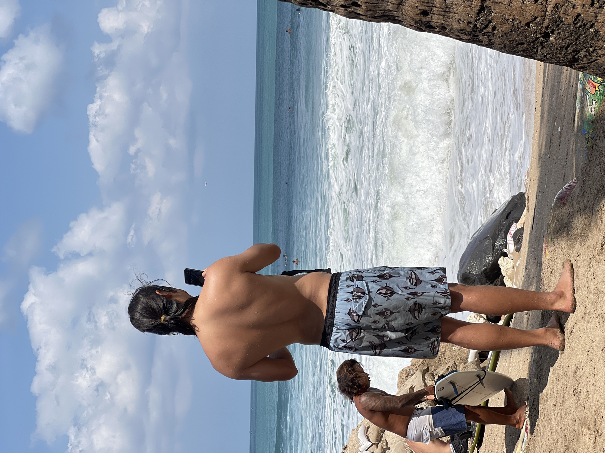 A man with dark hair tied in a ponytail stands on a sandy beach in Kuta, Indonesia, facing the ocean and holding a smartphone to take a photo. He is shirtless, wearing patterned swim trunks. To his left, another man with tattoos on his arm kneels, holding a surfboard. The ocean is visible in the background with waves breaking. Several other people are in the water. The sky is blue with scattered white clouds, and an airplane is visible in the distance. The foreground is sandy with some rocks and debris.