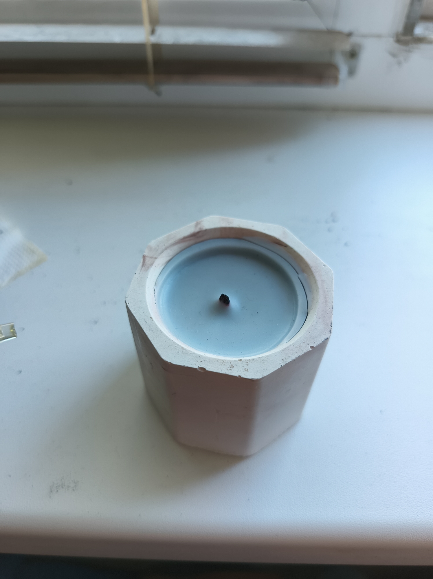 The image shows a pale blue candle in a hexagonal concrete holder. The candle holder is rough and unfinished, with a slightly uneven rim. The wax appears smooth and has a small, dark wick sticking out from the center. The candle is placed on a white surface, possibly a windowsill or table, which has some faint smudges and dust. In the background, there is a blurred horizontal bar above a lighter colored surface. To the left of the candle, a small white object with some texture, possibly a tissue, is visible. The overall lighting suggests an indoor environment, possibly a room with natural light coming from the side. The scene does not contain any identifiable landmarks or features that would indicate its location within Baranavichy, Belarus.