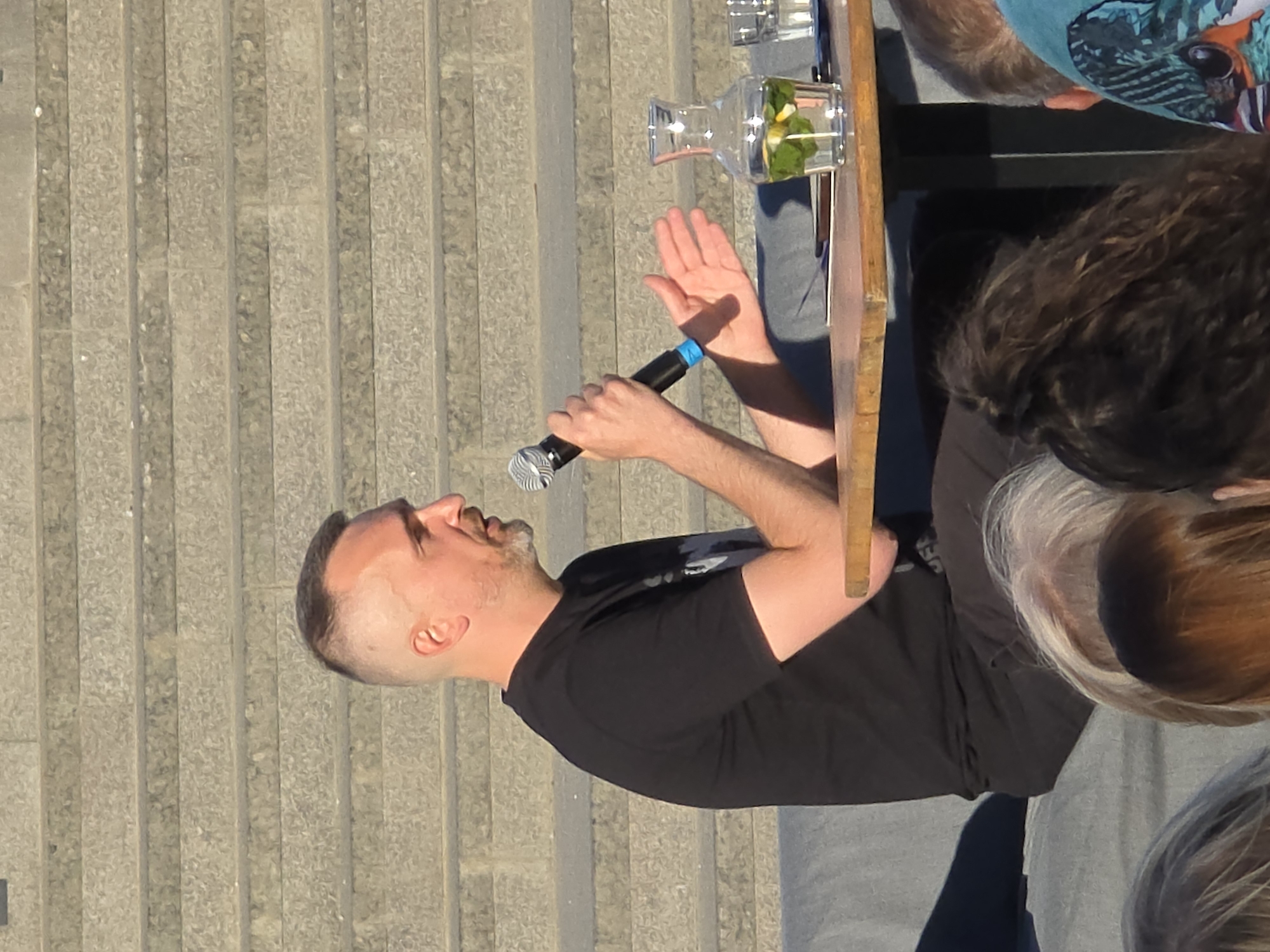 Captured from a high angle, the scene features a man speaking into a microphone at what appears to be a public event. The man, dressed in a black t-shirt, stands near a wooden table on which a pitcher of water with lemon slices and mint leaves sits next to empty glasses. The setting is outdoors, with a background of gray concrete steps. Other people are seated at the table, their faces obscured. The man is looking to his left with his hand raised. The image appears to have been taken during daylight hours.
The location is likely somewhere in Warsaw, Poland, given the cityscape and architectural details in the background. However, without more specific details, pinpointing the exact location within Warsaw is not possible.