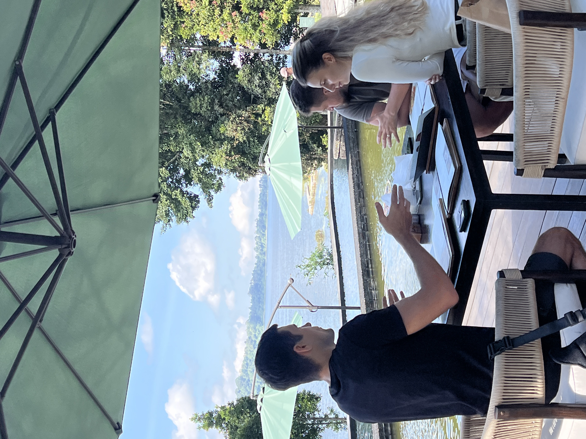 Three individuals are seated at a dark wood table on a wooden deck, situated outdoors. A large green umbrella provides shade over the table. In the foreground, a man wearing a black t-shirt is seen from the back, facing the other two individuals. He is gesturing with his hands as if speaking. To his right, a man wearing a grey t-shirt is visible from the side, looking towards the woman. Between them, a woman with long blonde hair, wearing a white long-sleeved top, is also visible from the side, looking forward. The table is set with menus and what appear to be papers and a phone. Beyond the deck, a body of water, possibly a lake, stretches out, with greenery and hills in the distance under a bright blue sky with scattered clouds. Two other green umbrellas are visible in the background, near the water. The scene appears to be a dining or meeting area at a lakeside resort or restaurant in Baturiti, Indonesia.