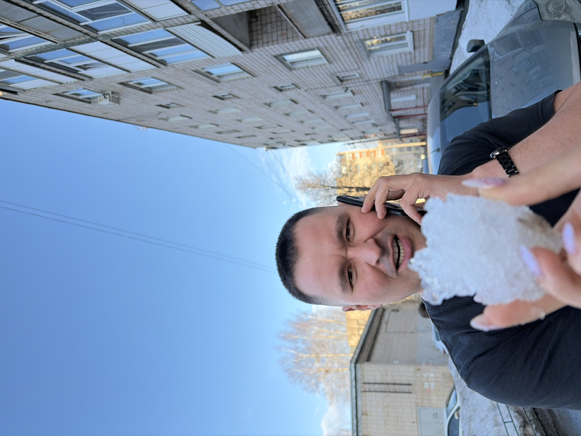 A man is holding a chunk of ice and talking on the phone, likely in Izhevsk, Russia, based on the typical Soviet-era architecture visible in the background. He is wearing a black t-shirt and a black watch. A woman's hand with manicured nails is partially visible near the ice. A multi-story brick building dominates the background, and there are bare trees and patches of snow on the ground. A gray car is also visible. The sky is clear and blue.