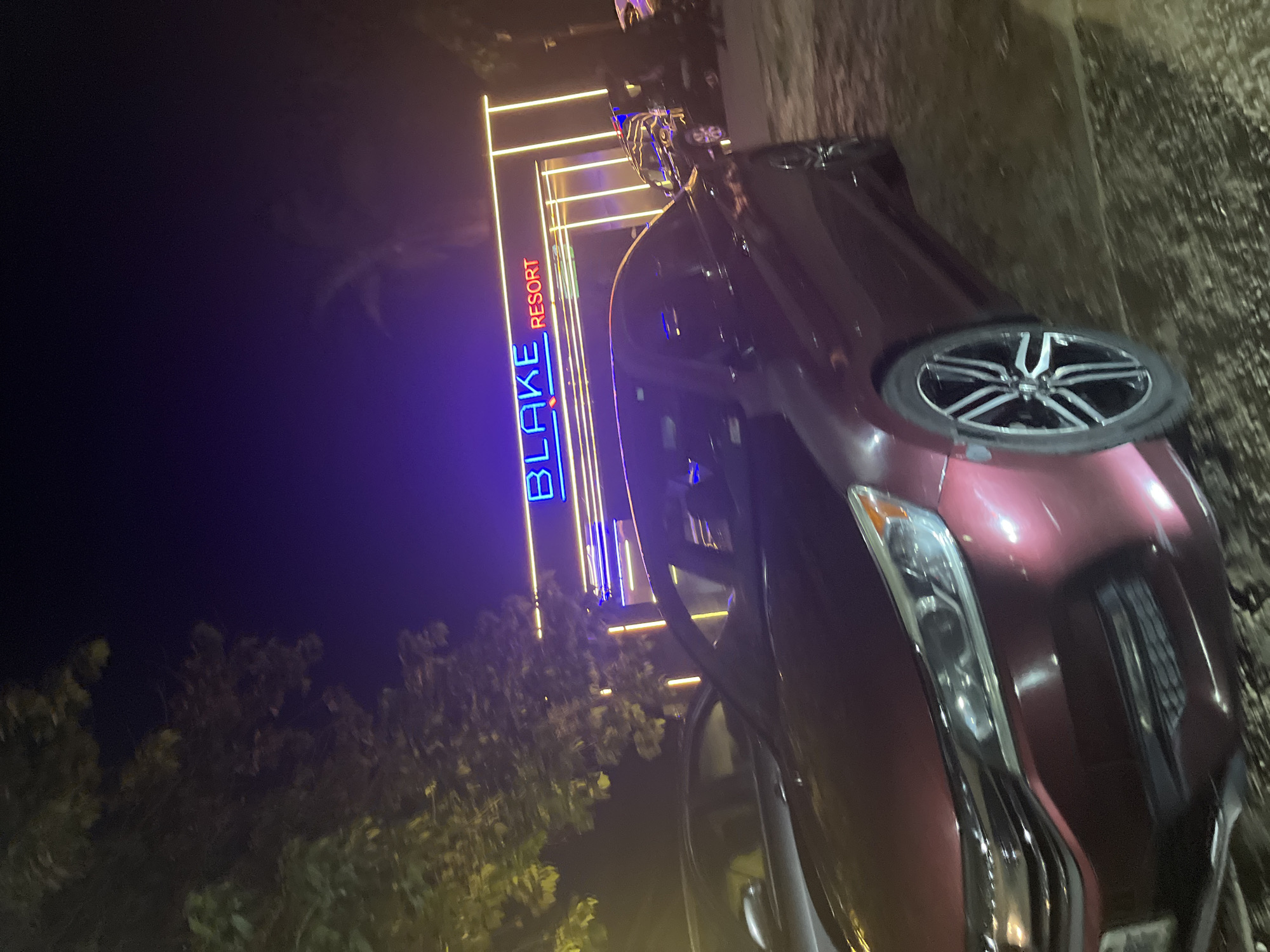 The image shows a maroon sedan parked in front of a building with a neon sign that reads "BLAKE RESORT". The building has a modern architectural design with vertical yellow lights. The scene appears to be at night, indicated by the dark sky and the illumination from the neon sign and building lights. Several other vehicles are partially visible in the background, also parked. Trees are present on the left side of the image, their leaves illuminated by the artificial lights. The foreground is dominated by the maroon car, with its headlights and front wheel clearly visible. The overall impression is that of an entrance to a resort or establishment at night. Based on the "BLAKE RESORT" signage, it is highly probable this location is in Abuja, Nigeria, as "Blake Resort" is a known establishment in that city.