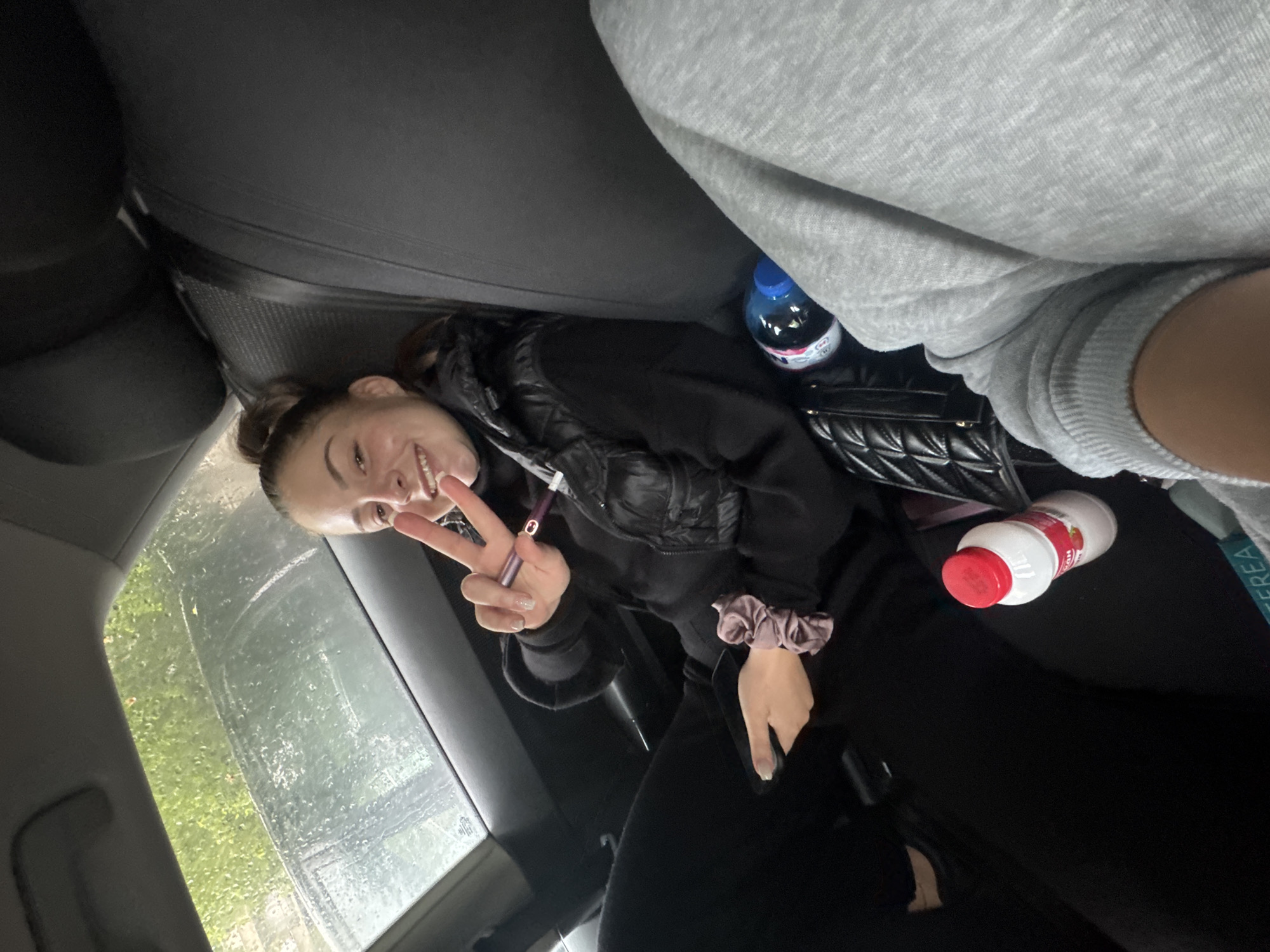 Inside what appears to be a car in Sofia, Bulgaria, a young person is captured in a casual, tilted perspective. The person, positioned in the back seat, is smiling broadly and giving a peace sign with their left hand. Their brown hair is pulled back, and they are wearing a dark hooded top layered with a black puffer vest. A light-colored scrunchie is on their right wrist.
In the foreground, part of another person wearing a grey ribbed sweatshirt is visible, suggesting a second occupant in the car. The setting is further defined by various objects in the car's interior: a black quilted handbag, a blue plastic bottle with "FRUTTIA" partially visible on its label, and a white plastic bottle with a red cap. The white bottle's label clearly shows "DANONE ACTIVIA," "Ягода" (Strawberry), and "МЮСЛИ" (Muesli), along with "НОВ СТАРТ НА ДЕНЯ" (New start of the day) in Bulgarian, indicating it's a yogurt drink. A dark purple cylindrical object, possibly a vape pen, is held in the main subject's hand.
Through the car window, raindrops are visible, suggesting it is raining or has recently rained. Green grass or foliage can be seen outside, indicating a daytime setting. The overall mood conveyed by the smiling subject and peace sign is one of happiness and relaxation.
