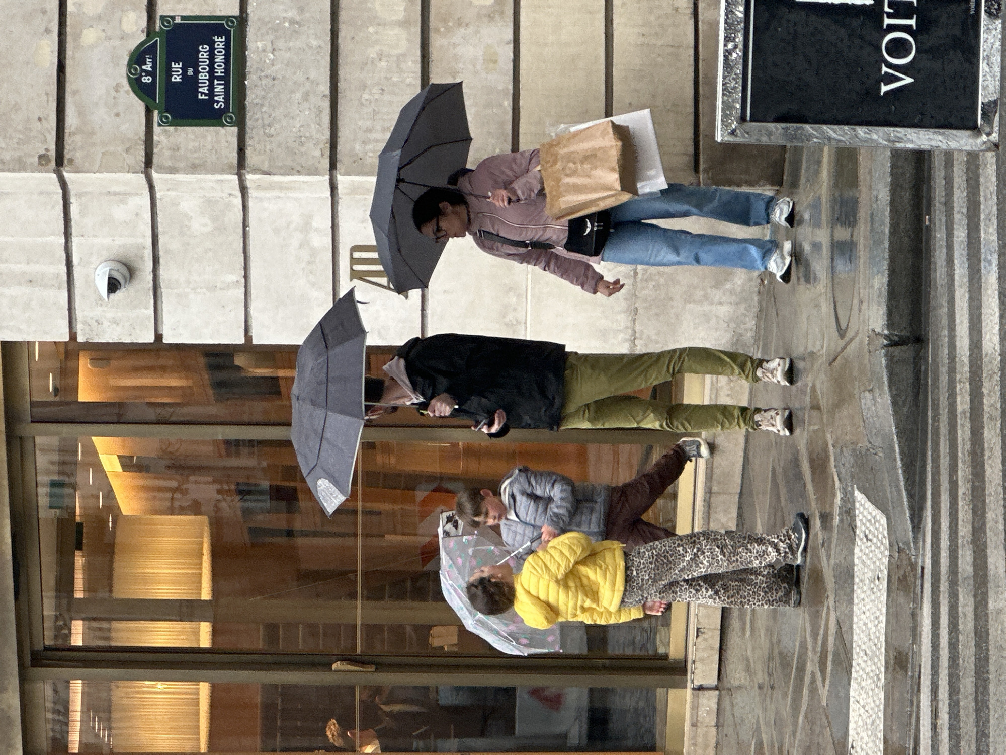The image captures a street scene in Paris, France, specifically on Rue du Faubourg Saint-Honoré. Four people are walking on the sidewalk in the rain, each holding an umbrella. The first person is wearing a pink jacket and blue jeans, carrying shopping bags. The second person is wearing a black jacket and green pants, using a phone. The third and fourth people, likely children, are walking together under one umbrella; one is wearing a grey jacket and the other a yellow jacket and animal print pants. The sidewalk is wet and reflects the surroundings. A street sign is visible on the upper left, indicating the location as "Rue du Faubourg Saint-Honoré." A store window is to the left, and a sign "VOIT" is on the right.
