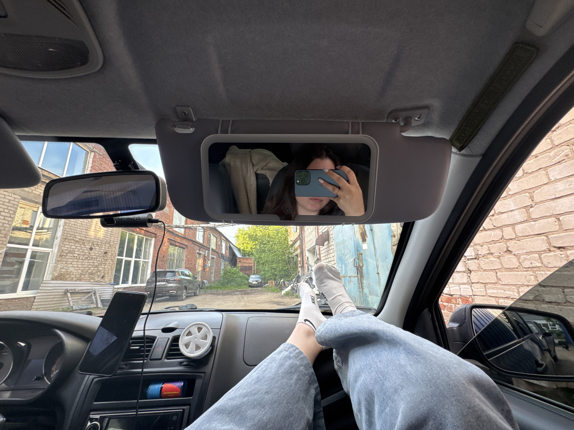 The image captures a first-person perspective from inside a parked car in Perm, Russia, looking out through the windshield. A person is sitting in the driver's seat, or possibly the passenger seat (judging by the reflection), with their feet propped up on the dashboard. The person's legs are clad in light-colored jeans, and they are wearing white socks, one of which clearly displays the "NIKE" logo.
The person is taking a selfie, visible in the car's sun visor mirror, which is flipped down. In the reflection, a woman with dark hair is holding a dark-colored smartphone, obscuring most of her face, with a ring visible on her finger. A light-colored garment is draped over the seat behind her in the reflection.
The car's interior features a dashboard with a smartphone mounted on a holder, a white circular air freshener, and a dash cam (DVR) attached near the rearview mirror. A charging cable runs from the dash cam. Below the central console, a small container, possibly an air freshener or cleaner, is partially visible, with text that appears to be "антидождь" (anti-rain). A green fabric strap with text, difficult to read but possibly English, is attached to the ceiling lining.
Outside the car, an urban or semi-urban environment is visible. Brick buildings with multiple windows line a narrow street or alley. A dark-colored SUV is parked in the mid-ground, and another car is visible further down the road, partially obscured by trees. A large blue metal garage door is seen on the right. The external rearview mirror on the right shows a reflection of a brick wall and potentially a building.
The scene takes place during daytime, likely late afternoon, indicated by the natural light and shadows. The sky appears clear. The overall mood is relaxed and casual, as suggested by the person's posture.