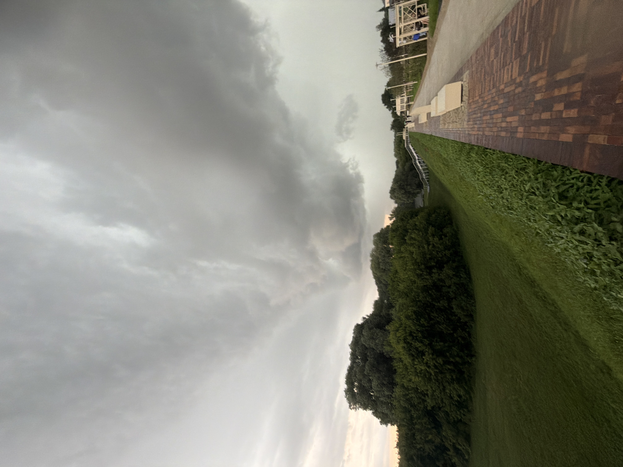 The image depicts a view of the Kolomna Kremlin embankment in Kolomna, Russia, under a heavily overcast sky with dramatic, dark clouds. A paved walkway, constructed with reddish-brown bricks and lighter concrete sections, runs diagonally across the lower right portion of the frame. Several light-colored, minimalist benches are positioned along the walkway. To the left of the walkway, a steep, grassy slope covered in lush green grass and vegetation descends. Beyond the slope, a dense line of dark green trees forms a natural border. In the distance, along the walkway, a few figures can be seen walking. Further in the background, white structures resembling gazebos or open pavilions are visible, with some people sitting or standing near them. The overall atmosphere is subdued, likely due to the impending or ongoing bad weather indicated by the prominent storm clouds.