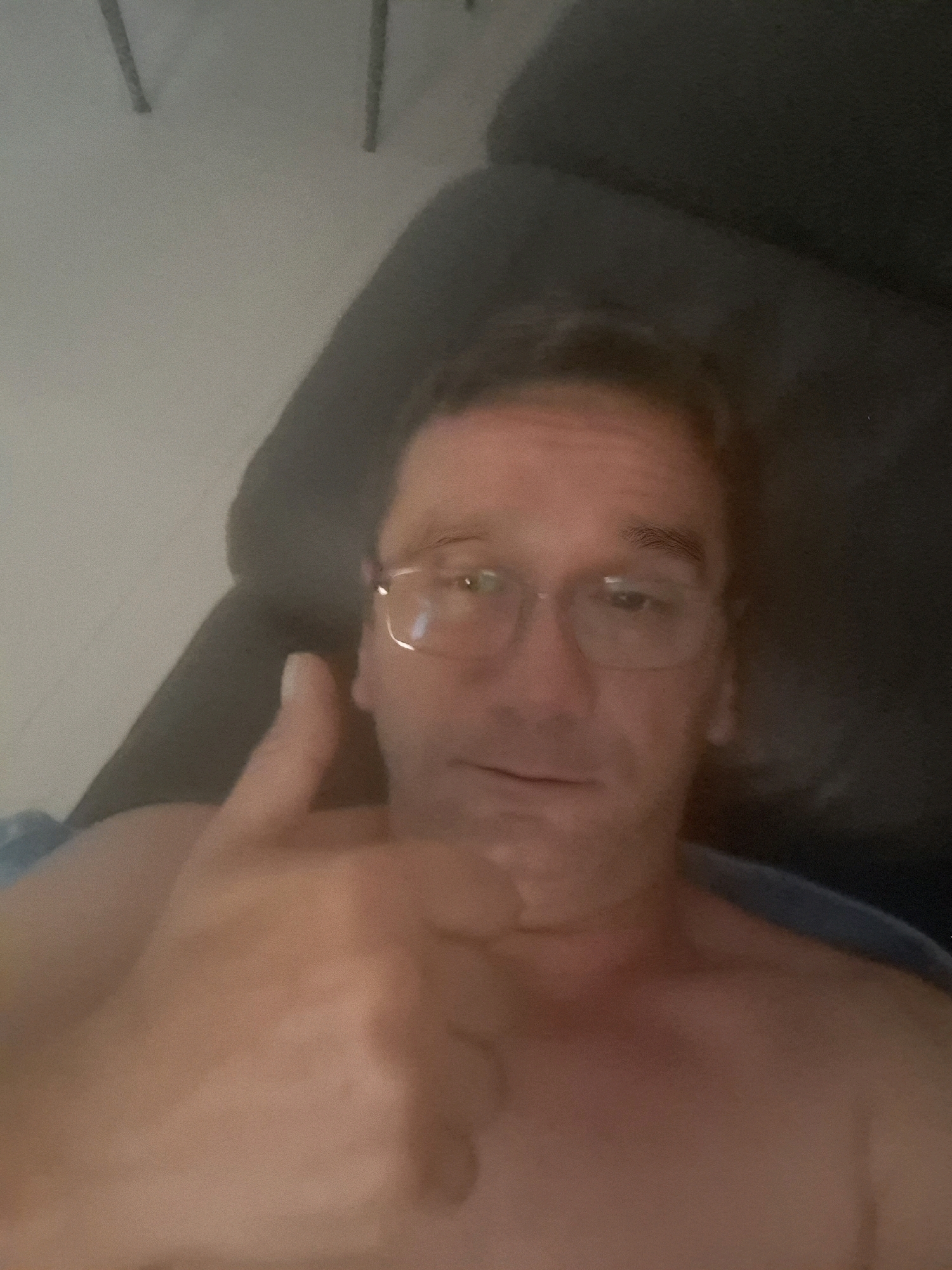 A selfie taken indoors shows a man with glasses giving a thumbs up, lying down with his chest visible. The background is dark and indistinct. The image was taken in Yerocham, Israel.