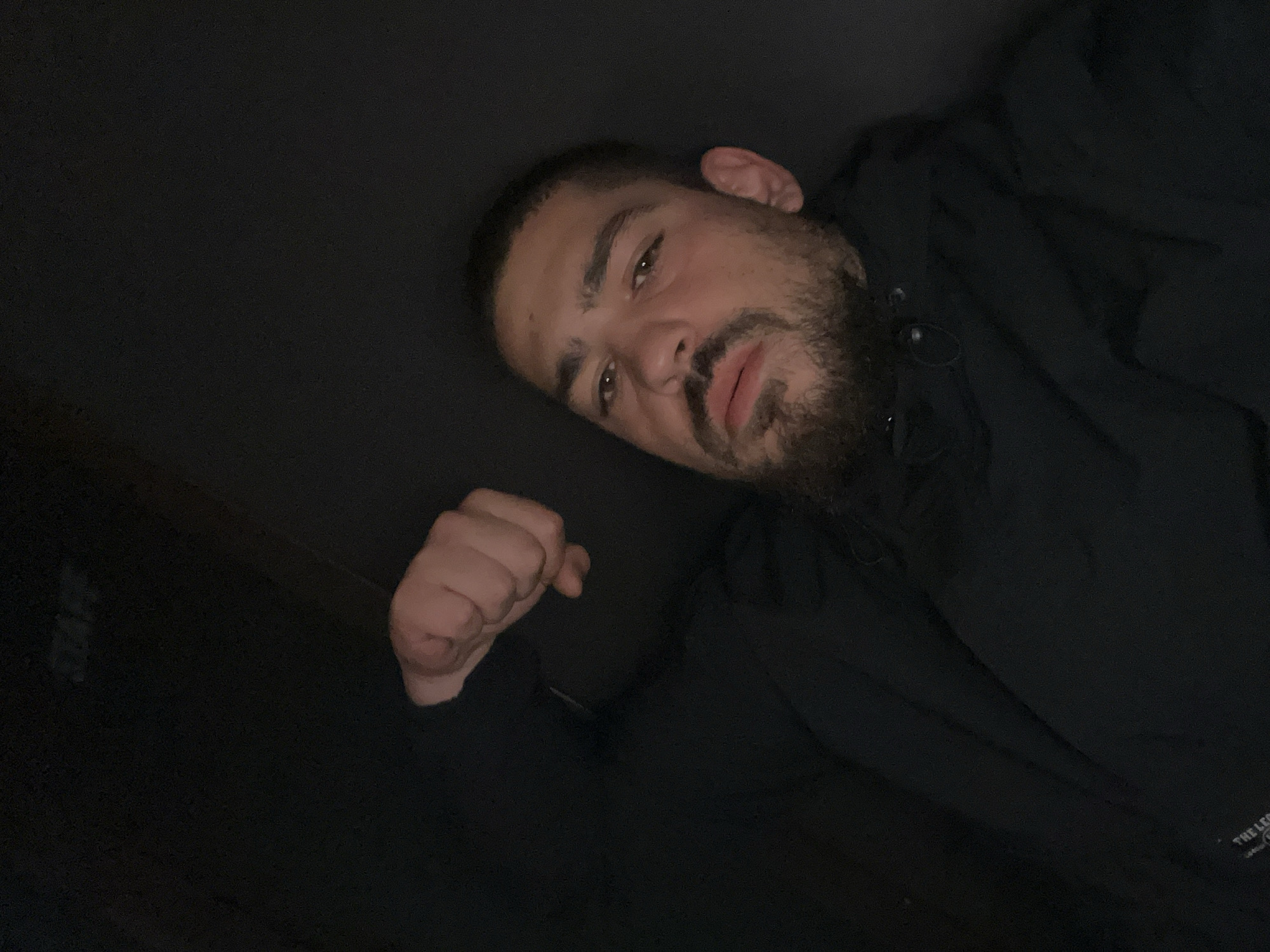 A close-up, dimly lit selfie shows a man from the chest up. He has short dark hair, brown eyes, a mustache, and a beard. He is wearing a black hooded jacket, and his left fist is raised towards the top left of the frame. The background is dark and indistinct. There is a label on the lower left of the jacket that reads "THE LEG". No specific location in Burgas, Bulgaria, is identifiable.